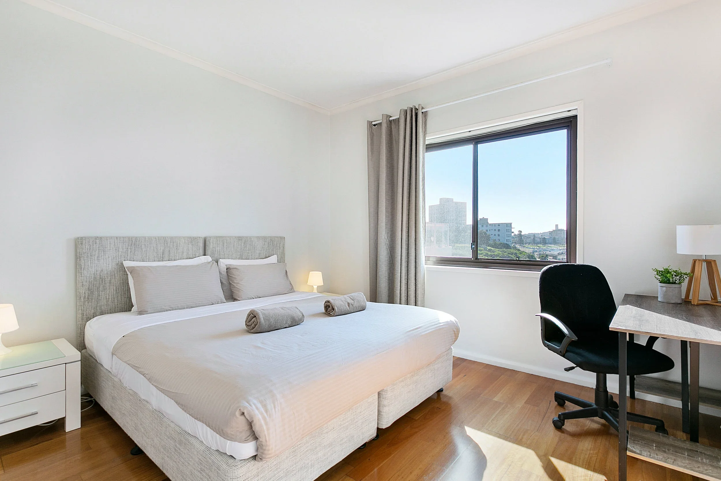 A bedroom with a bed, pillows, bedside lamps, a window with curtains, a desk with a chair, a lamp, and a potted plant, cityscape view outside.