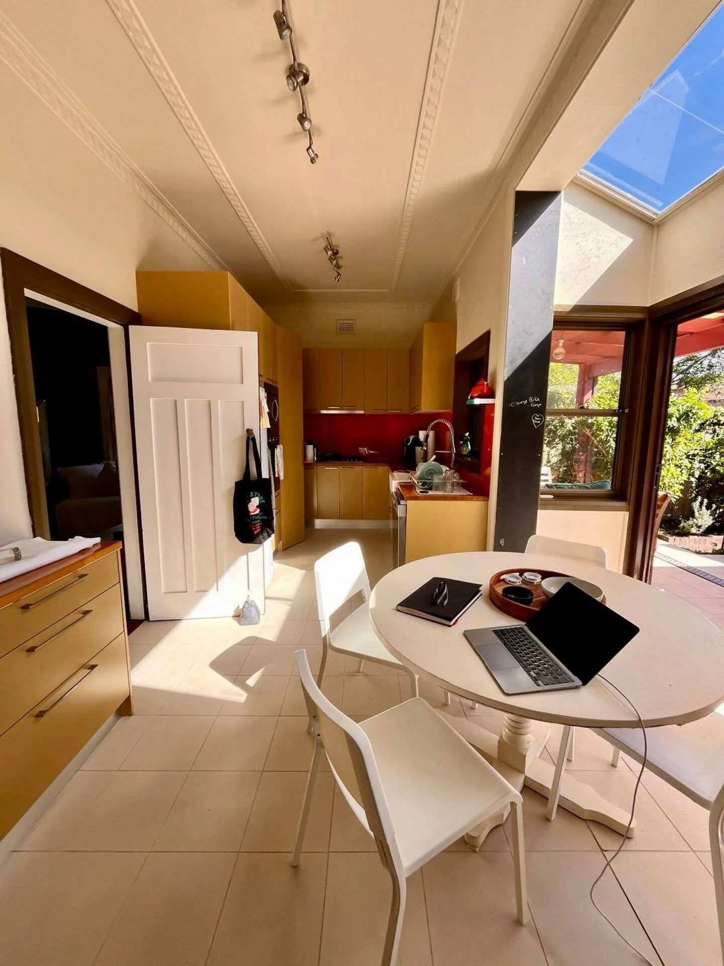A sunlit kitchen and dining area with a round white table, three white chairs, a laptop, notebook, and other items. There is a large window and sliding door leading to an outdoor patio with trees. The ceiling has track lighting, and the kitchen has w