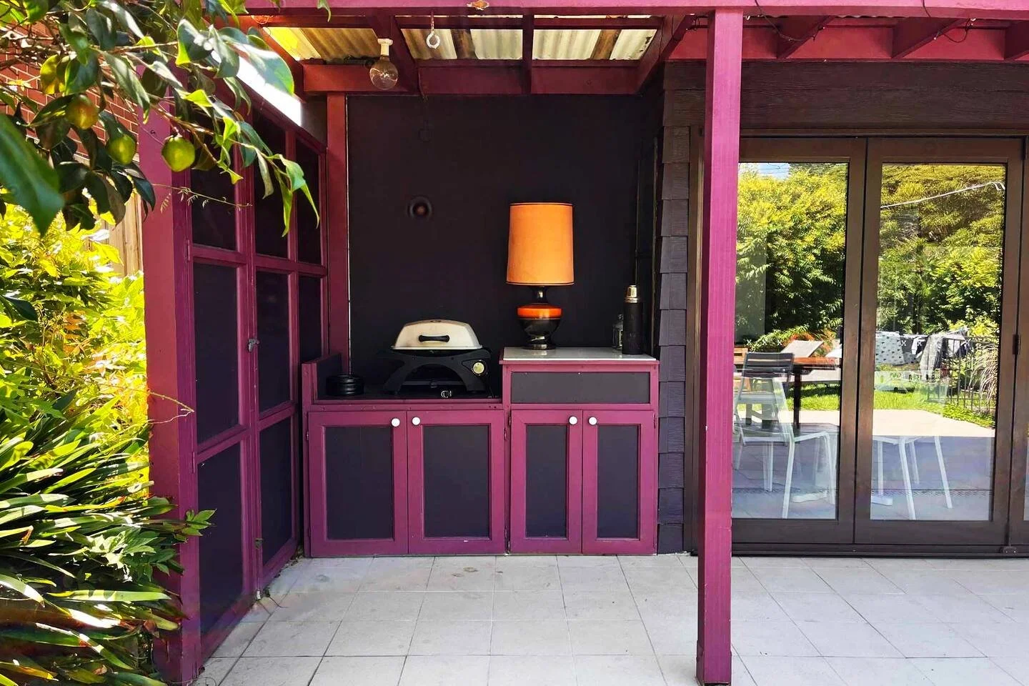 Outdoor patio area with purple cabinets, a lamp, a grill, and a sliding glass door revealing a backyard with outdoor furniture and green trees.
