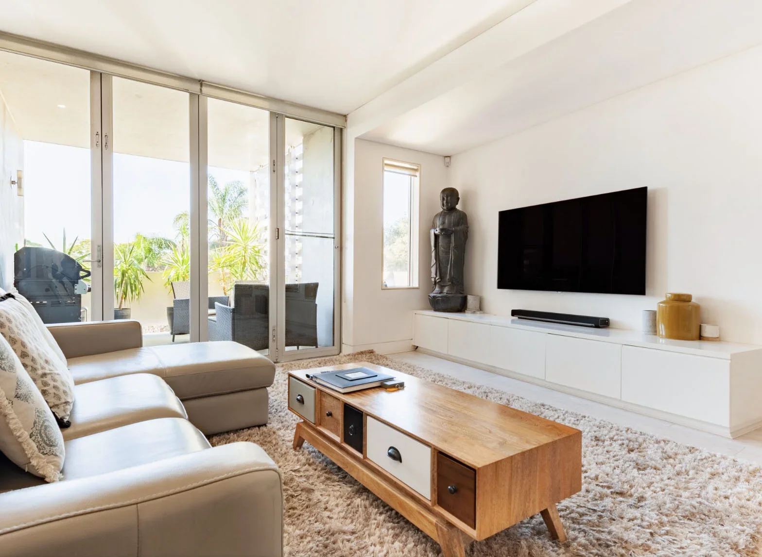 Modern living room with beige sofa, wooden coffee table, wall-mounted TV, sculpture, and an outdoor patio with chairs and potted plants.
