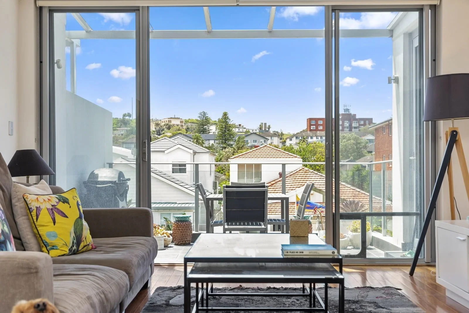 Living room with a large sliding glass door opening to a balcony, showing rooftops and trees outside on a sunny day.