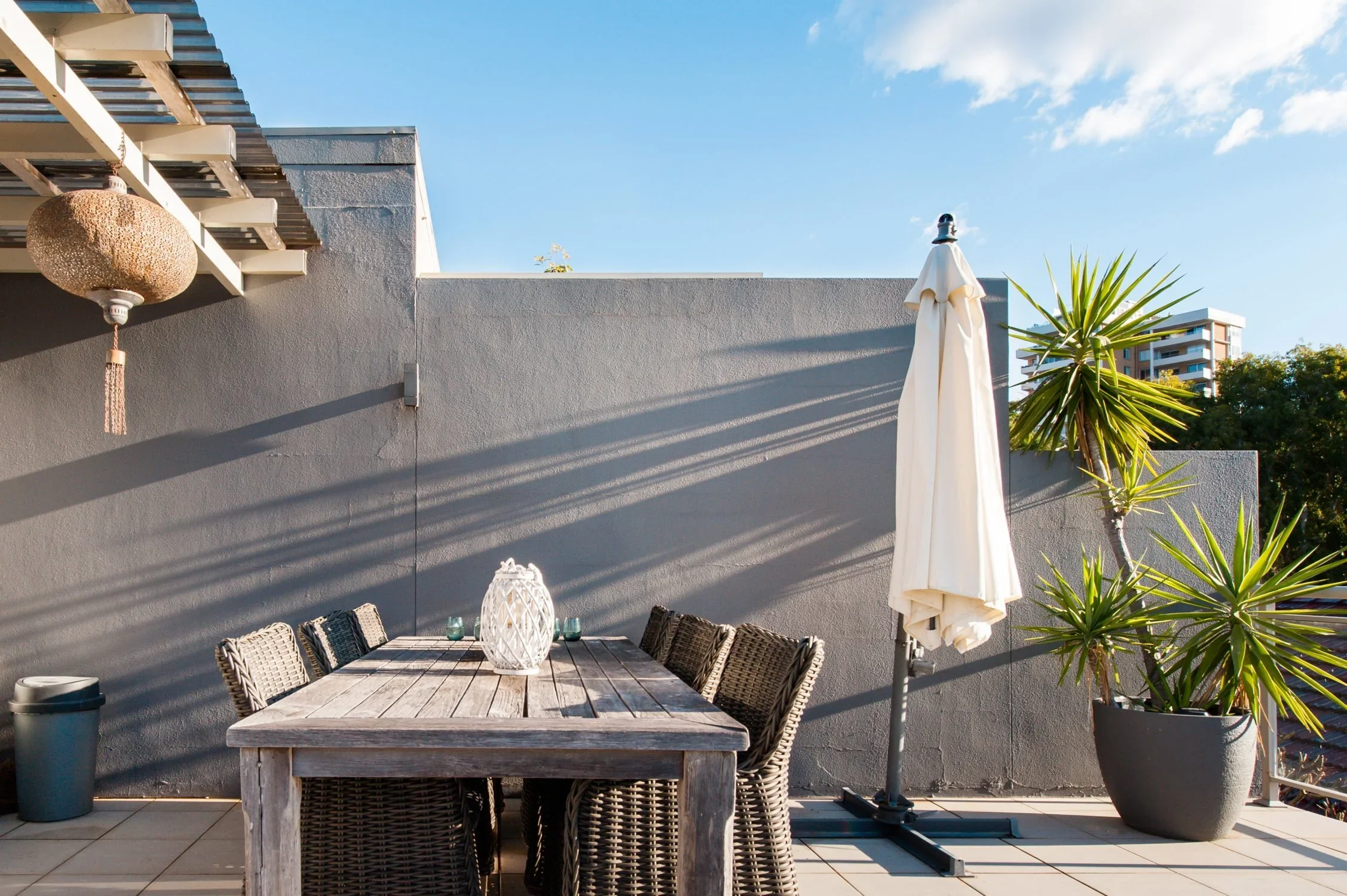 Outdoor patio with a wooden table and wicker chairs, a closed white patio umbrella, large potted plants, a wall-mounted lamp, and a clear blue sky.