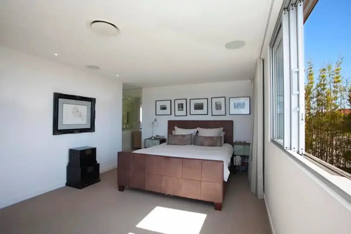 A bedroom with a large bed, framed pictures on the wall, a window open to the outside with trees, a small black TV stand, and a mounted TV.