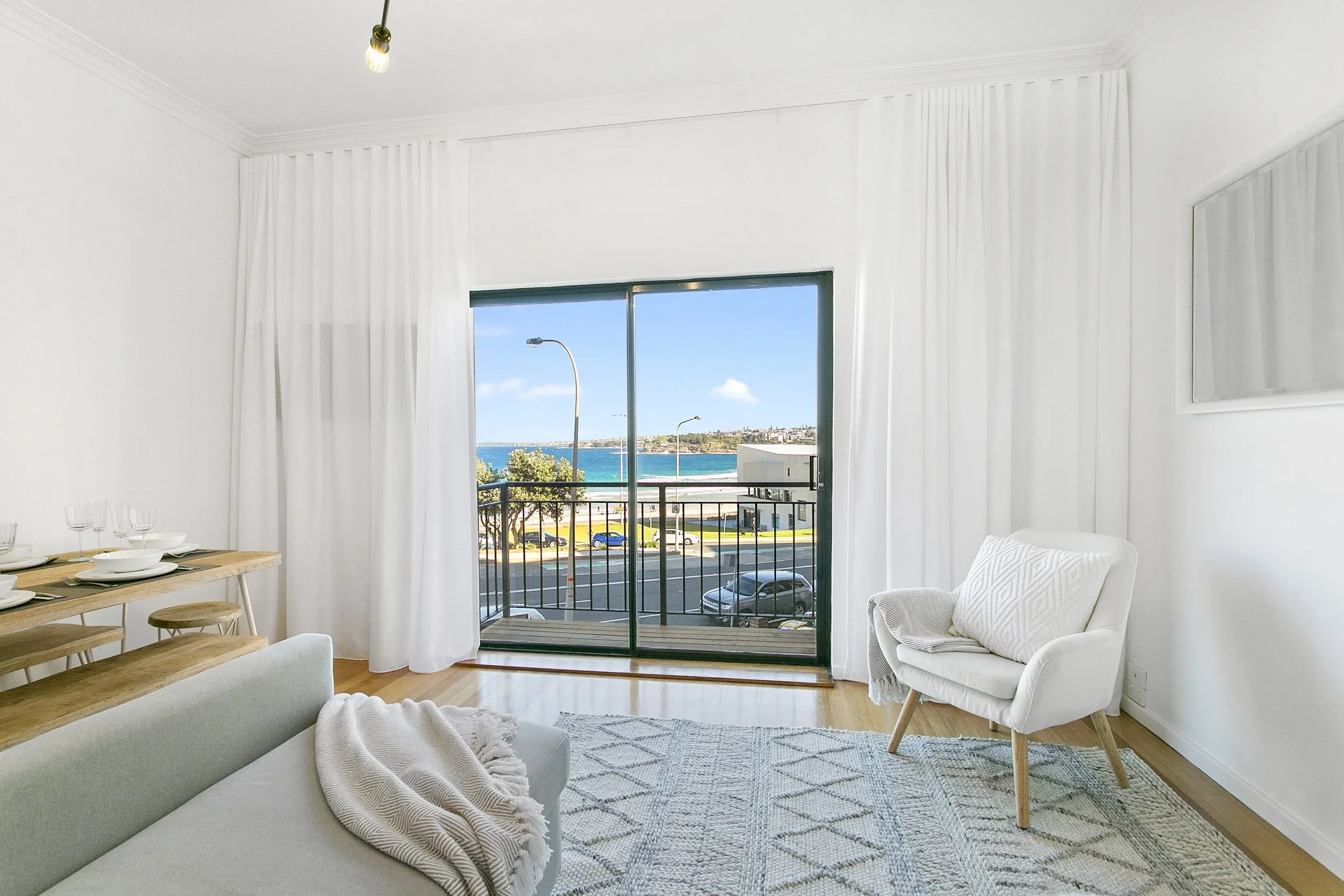 Living room with white walls, a sliding glass door leading to a balcony, and a view of the ocean with street lamps and cars outside. The room has white curtains, a white armchair, a light-colored rug, and a section of a dining table with place settin