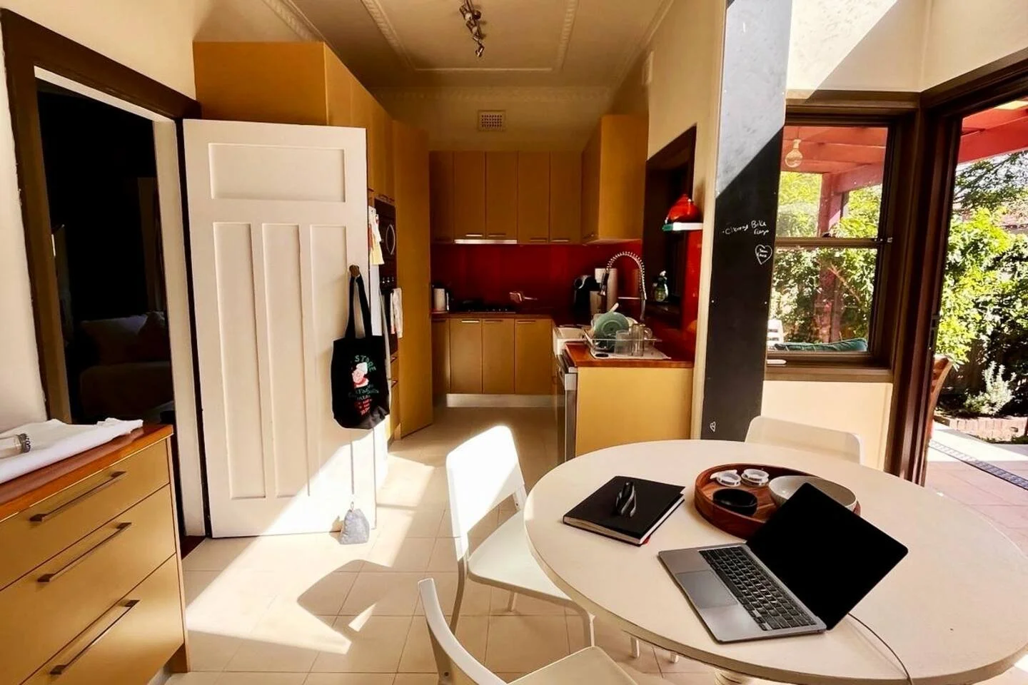 Kitchen with a round dining table, opened window showing outdoor patio, and white door with black bag hanging on it.
