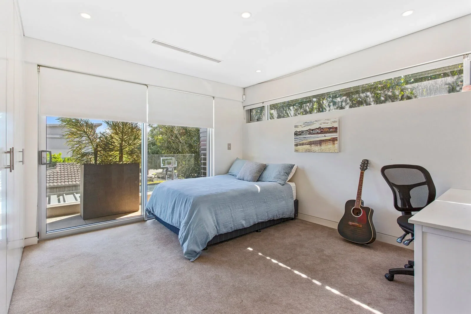 Bedroom with a bed, guitar, desk, office chair, and large windows with a balcony and outdoor view