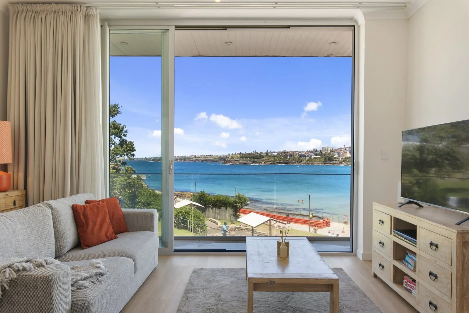 Living room with large glass window view of a blue ocean, beach, and sky, with living room furniture including a beige sofa with orange pillows, a wooden coffee table, a TV on a wooden stand, and light-colored curtains.