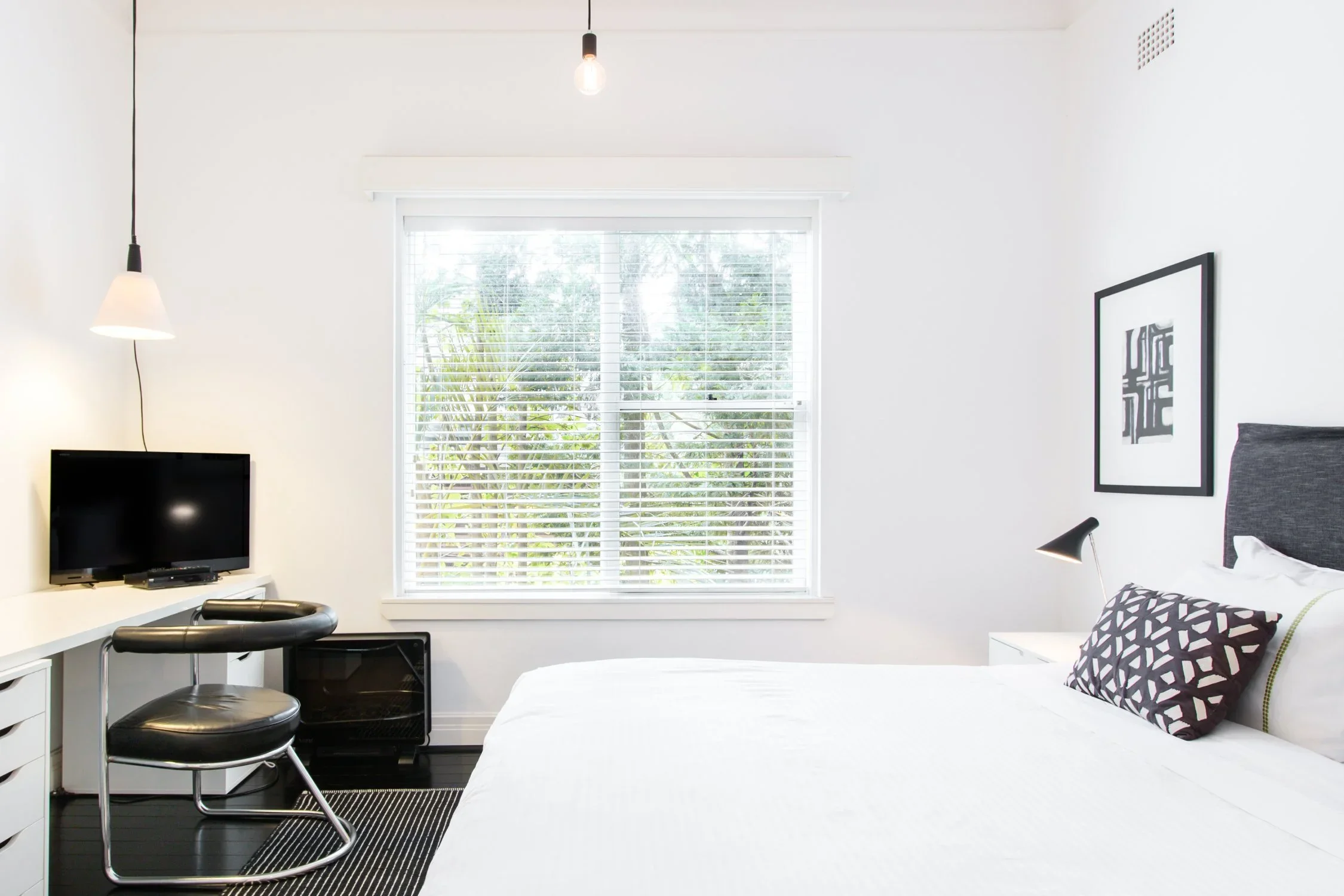 A bright, minimalist bedroom with white walls, a large window with white blinds, a bed with white bedding and a decorative black and white pillow, a black framed abstract art on the wall, a small bedside lamp, and a white desk with a computer monitor