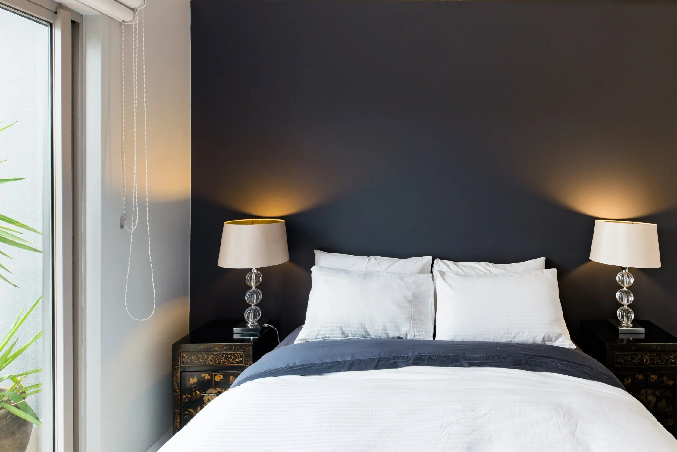 A neatly made bed with white pillows and bedding, flanked by two black nightstands with decorative lamps, set against a dark gray accent wall, with a window partially visible to the left.