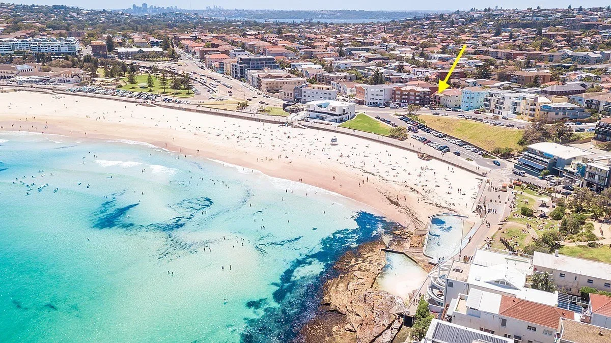Aerial view of a beach with turquoise water and sandy shoreline, adjacent to a cityscape with residential and commercial buildings, parking lots, and green parks.