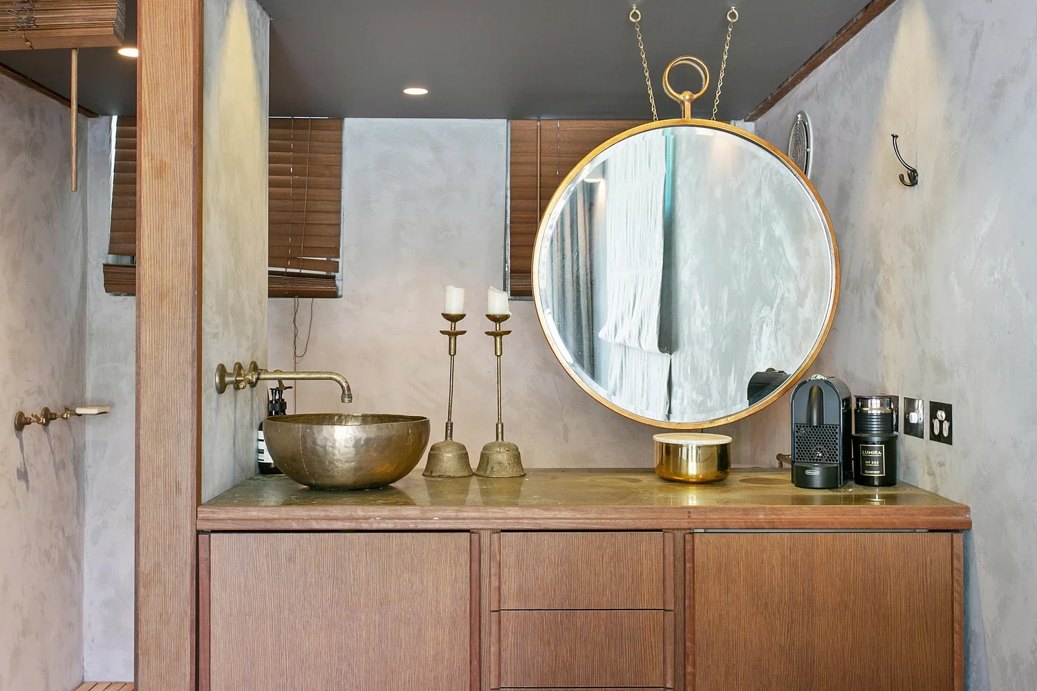 A bathroom vanity with a brass sink, two candlesticks, a round mirror, a coffee machine, and decorative items in an elegant, modern setting.
