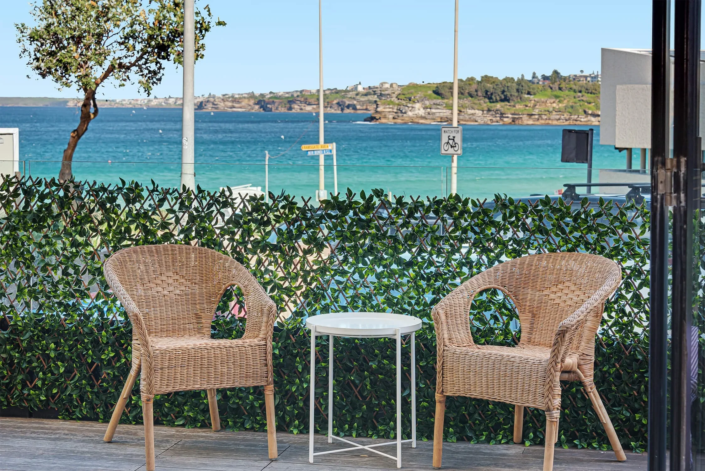 Two wicker chairs with a small white round table between them on a deck, overlooking a body of water with an island in the distance and street signs visible in the background.