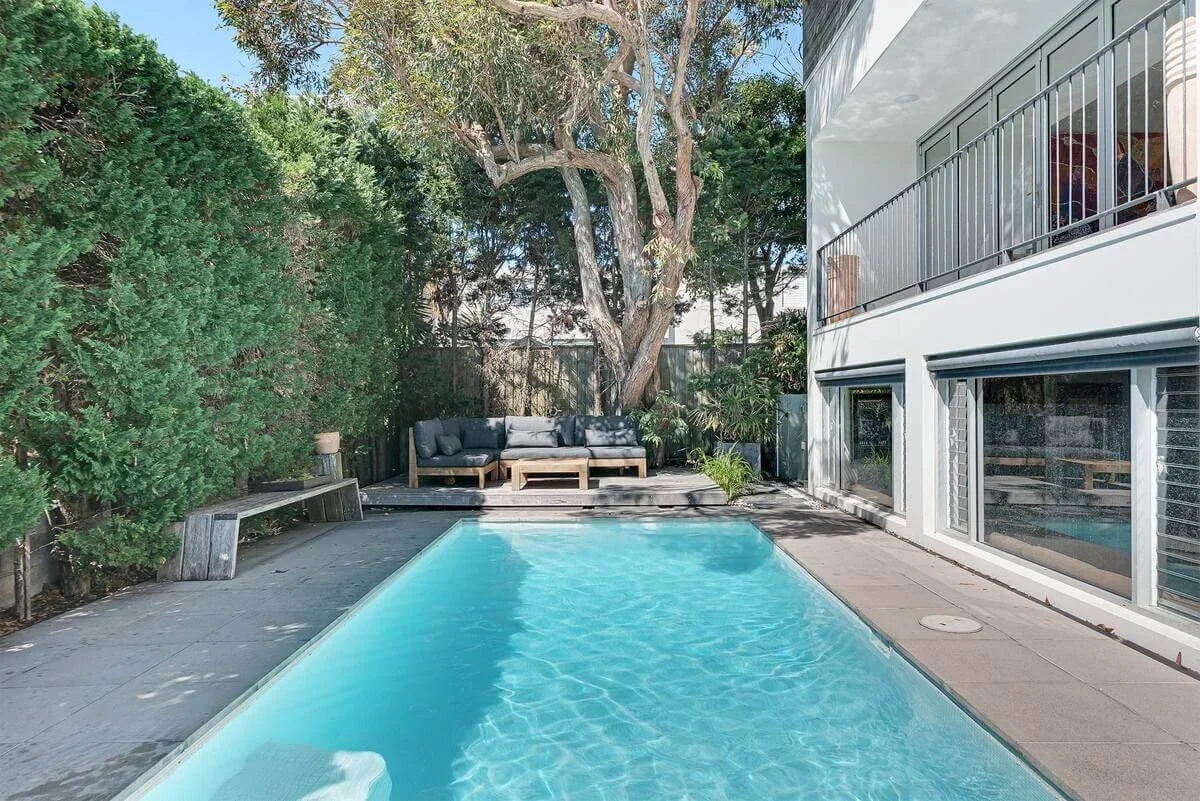 Residential backyard with a swimming pool, outdoor seating area under a tree, and a modern house with large windows and a balcony.