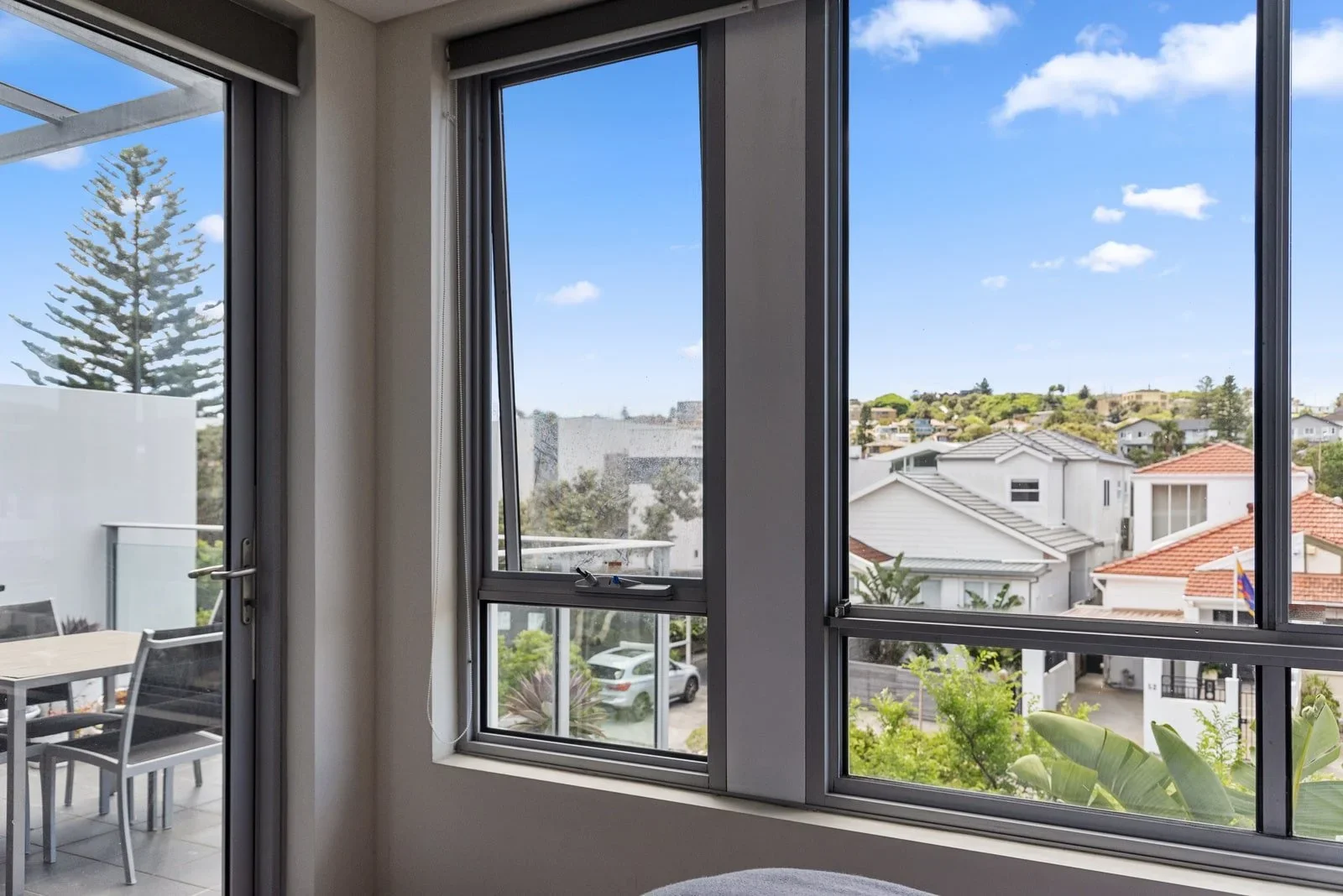 View from inside a room showing open sliding glass windows overlooking a neighborhood with houses, trees, and a partly cloudy sky.