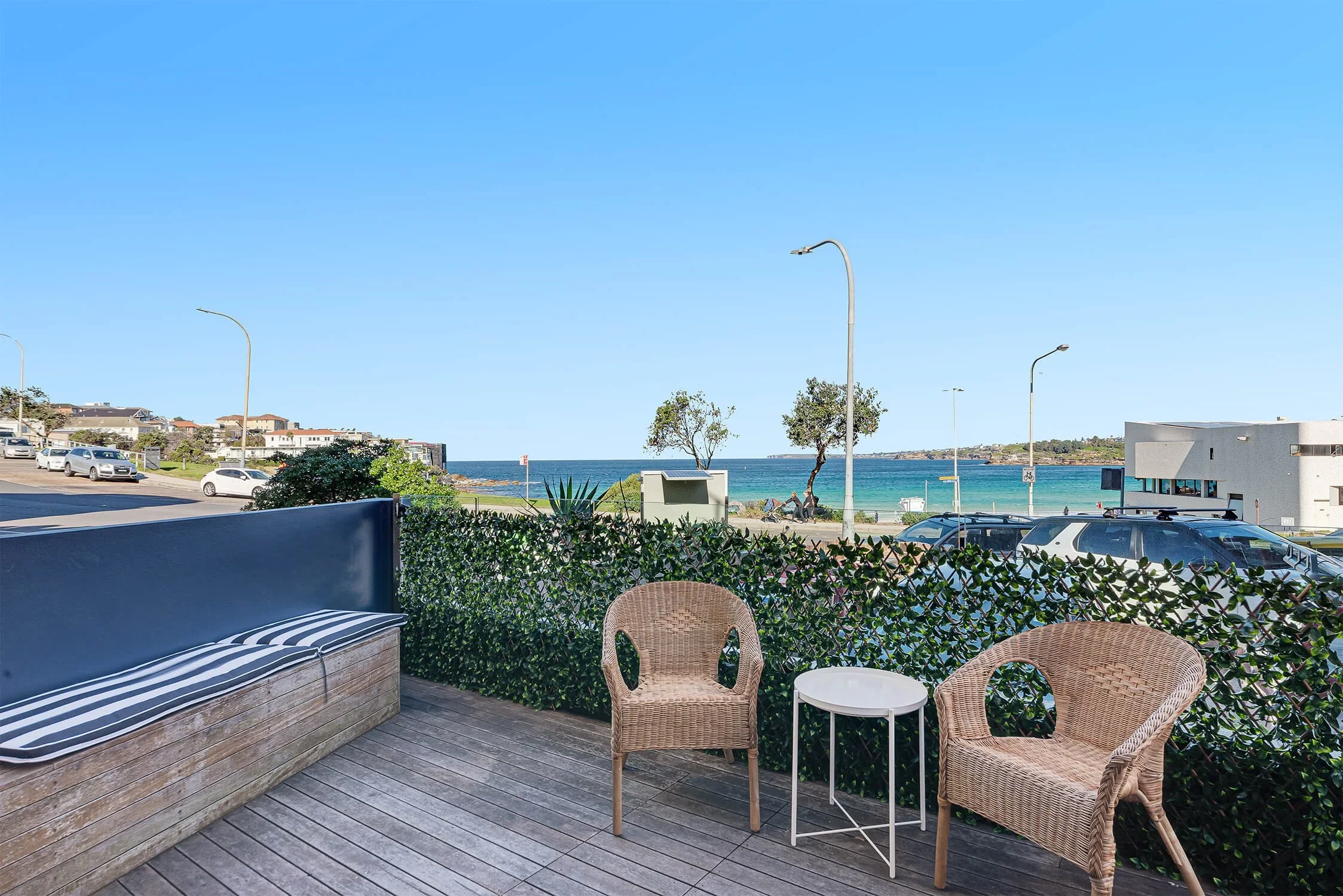 A seaside patio with two wicker chairs and a white side table, overlooking a parking lot, a leafy hedge, and a beach with the ocean in the background under a clear blue sky.