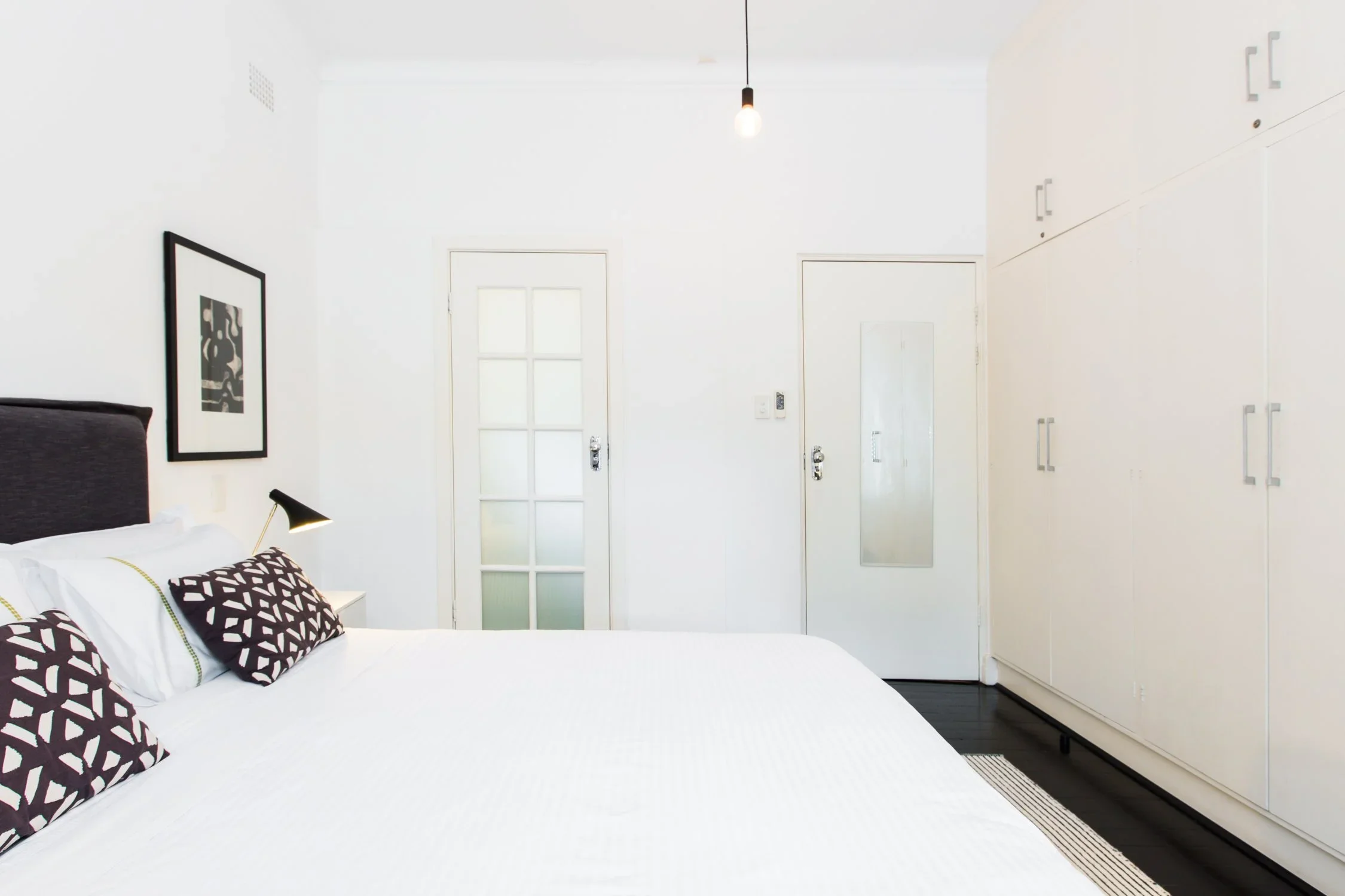 A minimalist bedroom with white walls, a bed with black and white pillows, a black bedside lamp, artwork on the wall, a white door, a mirror, a large white wardrobe, and a black and white rug.