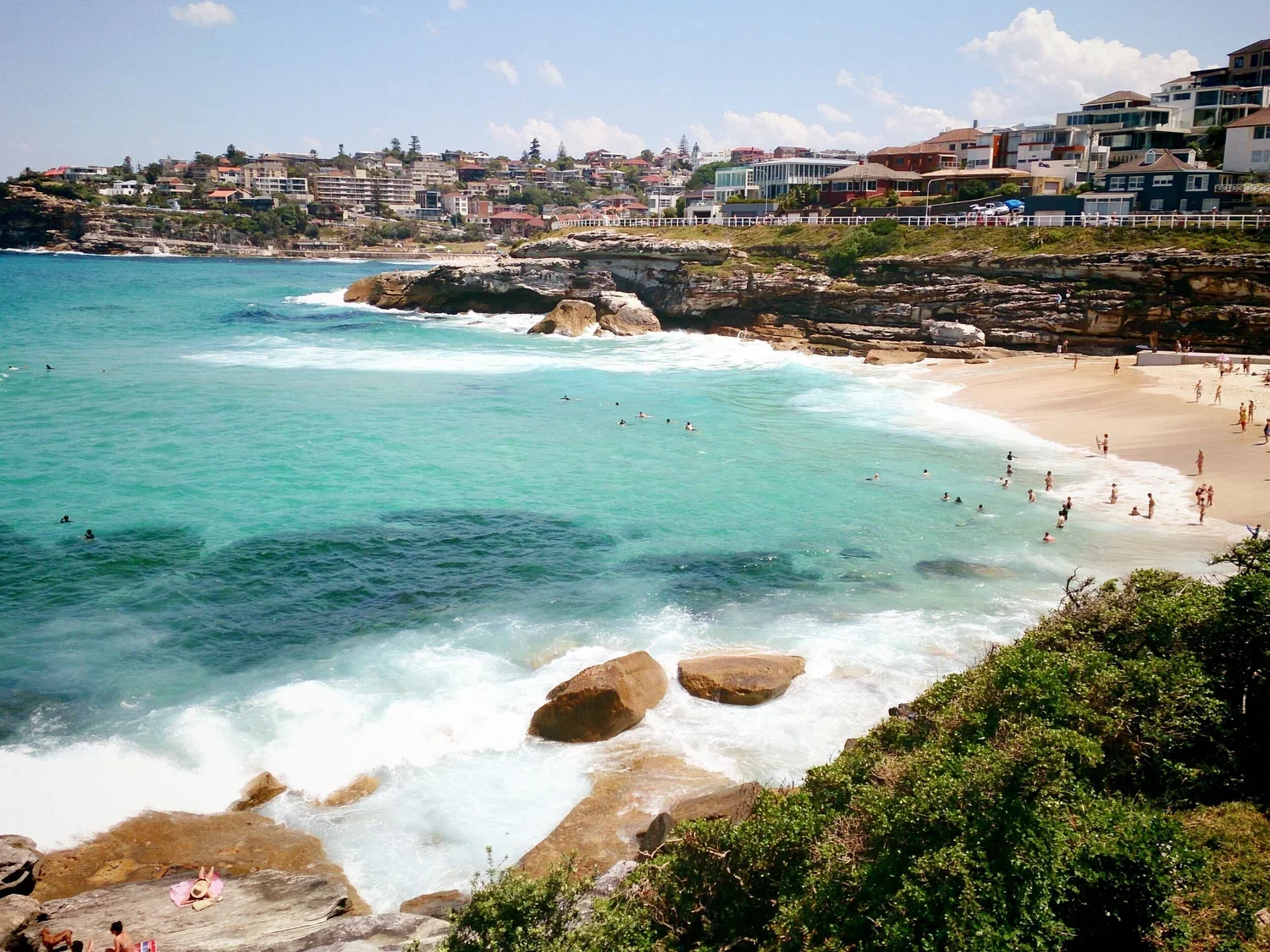 View of a beach with turquoise water, sandy shore, and rocks in the foreground. People are swimming and relaxing on the beach. Residential buildings are on the hillside in the background, under a cloudy sky.