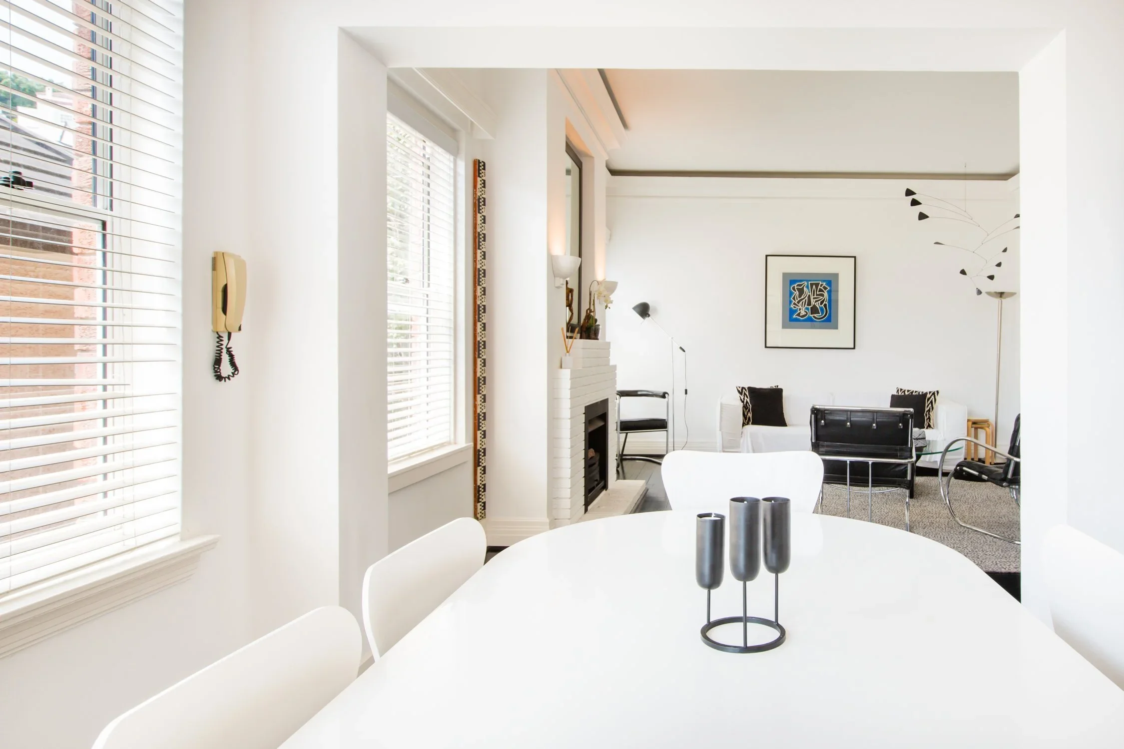 Modern living room and dining area with white walls and furniture, black and white artwork, and large windows with blinds.
