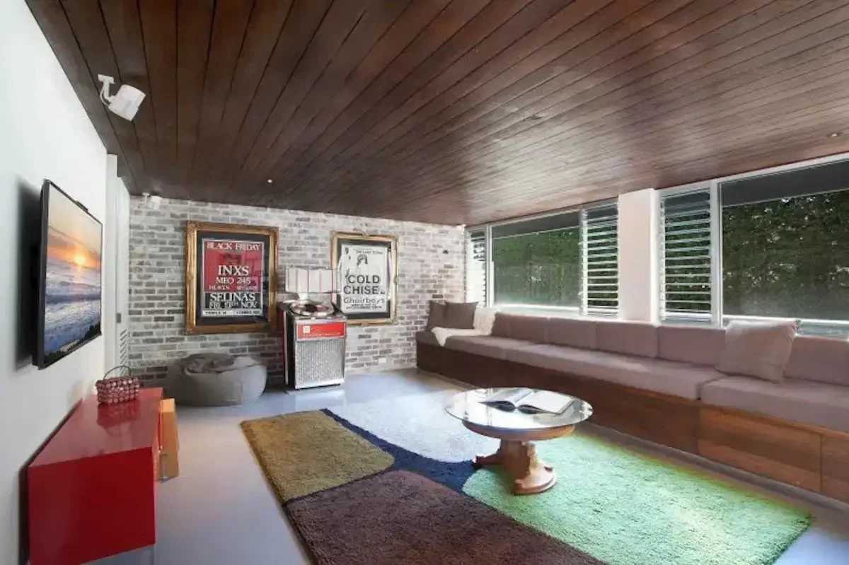 Living room with a mounted flat-screen TV, a multicolored rug, a long beige couch along the windows, and framed posters on the wall.