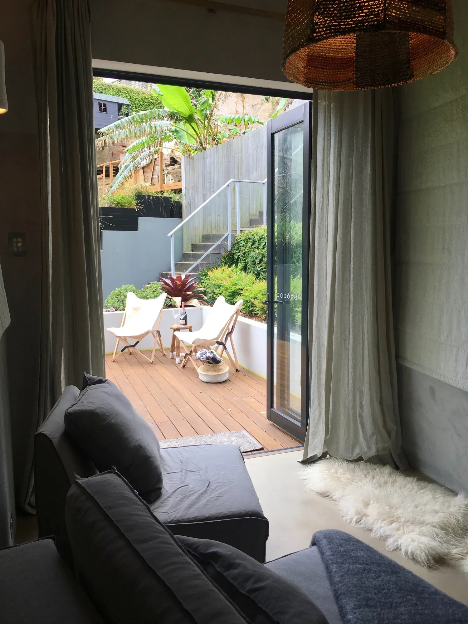 Living room with an open sliding glass door leading to an outdoor patio. The patio has two white deck chairs, a small table with a wine bottle, and greenery including large banana leaves and bushes. The indoor area has dark sofas, a white shaggy rug,