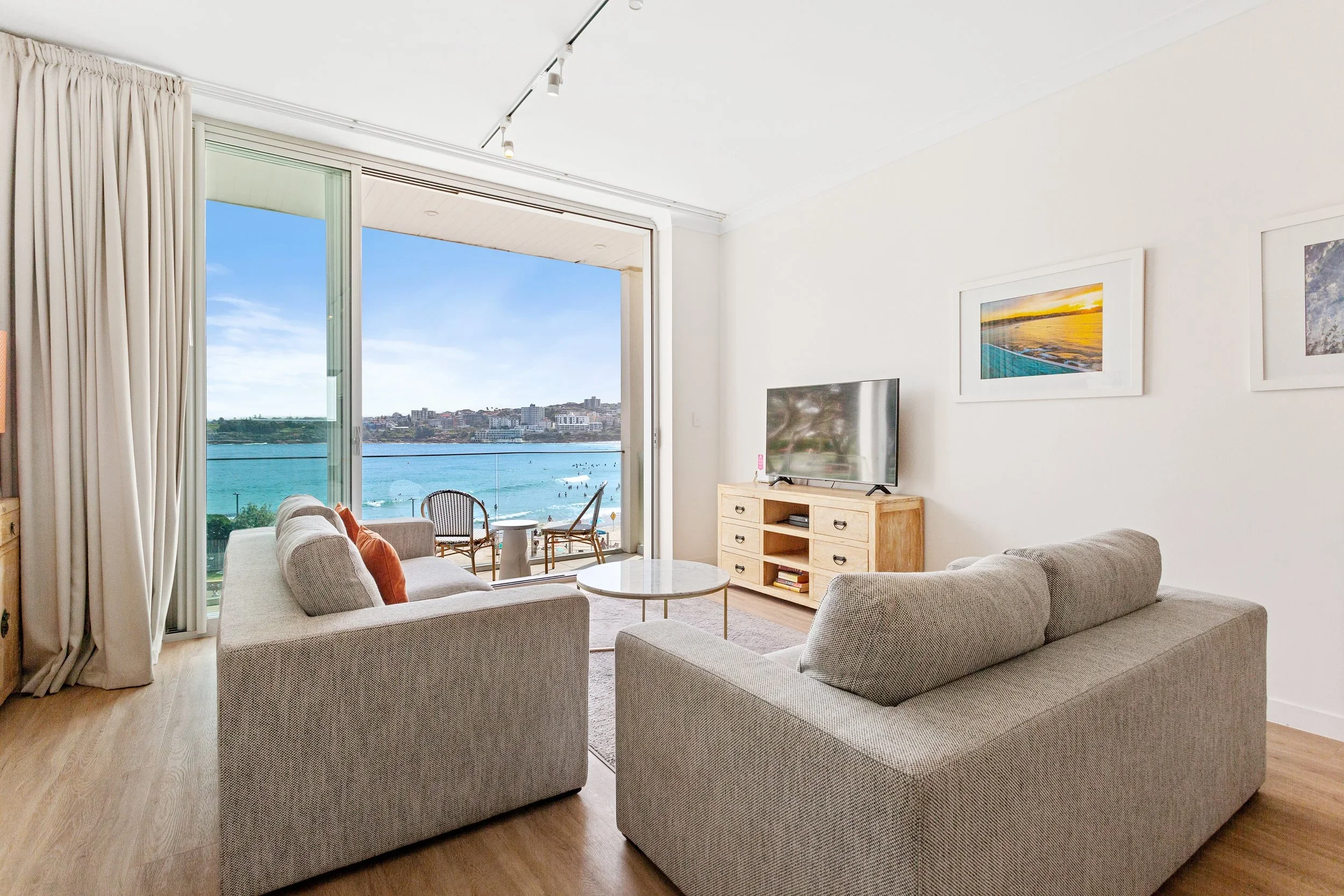 Living room with two gray sofas, a wooden TV stand with a flat-screen TV, a coffee table, and balcony door opening to a view of blue water and city skyline.