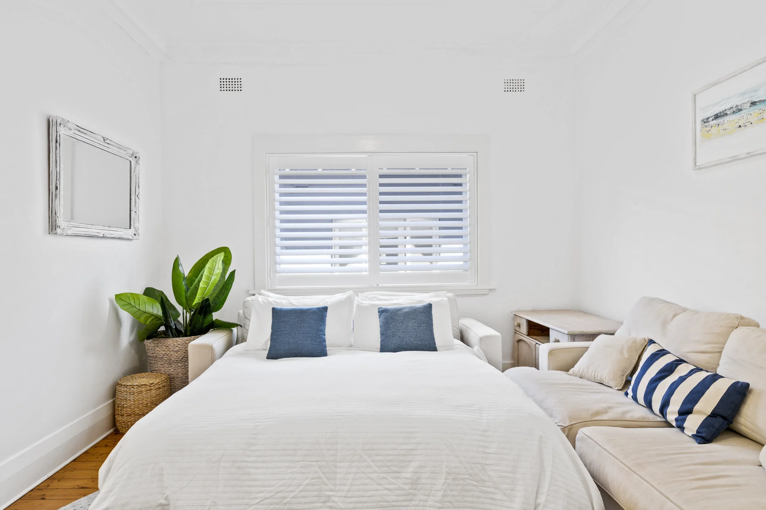 Bright, minimalistic bedroom with a white bed, blue pillows, a beige sofa with striped and plain pillows, a potted plant, and framed artwork, with a window that has horizontal blinds.
