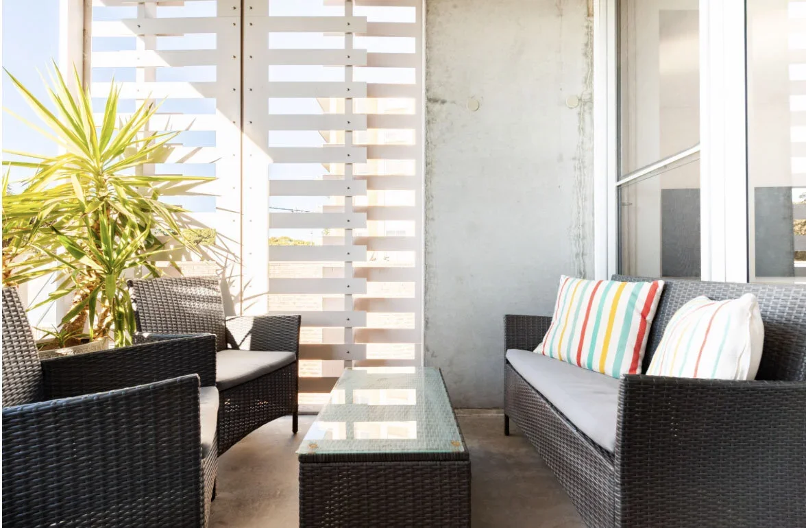 Outdoor balcony seating area with black wicker furniture, including a loveseat and two armchairs, with striped pillows, a glass-top table, and a large green plant, surrounded by white wooden slats and concrete walls.