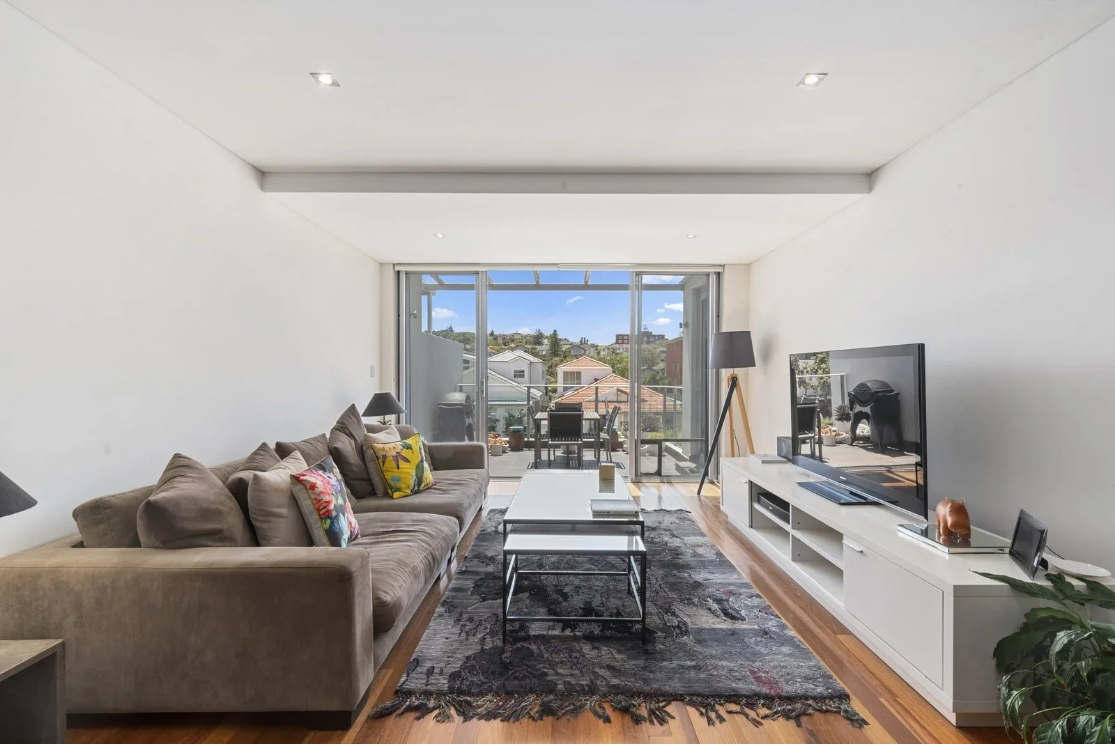 Modern living room with beige sofa, black lamps, and colorful cushions, white entertainment unit with TV, large glass sliding doors leading to balcony with outdoor furniture, hardwood floors, and a patterned area rug.