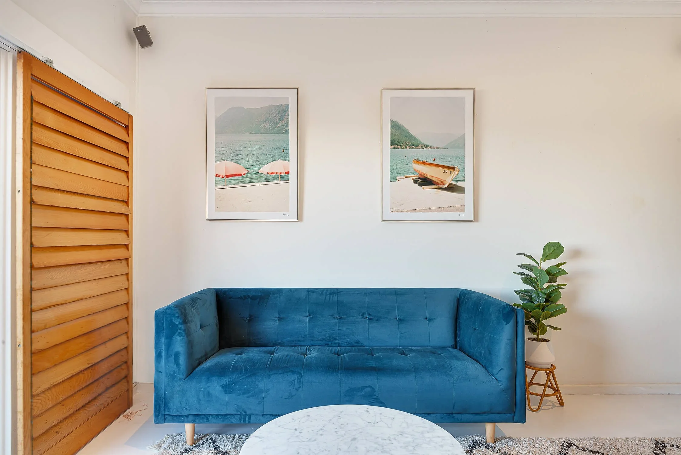 Living room with a blue velvet sofa, a potted plant in a white pot on a wooden stand, two framed pictures of a lake and mountains on the wall, a round marble coffee table, and a wooden sliding door with horizontal slats.