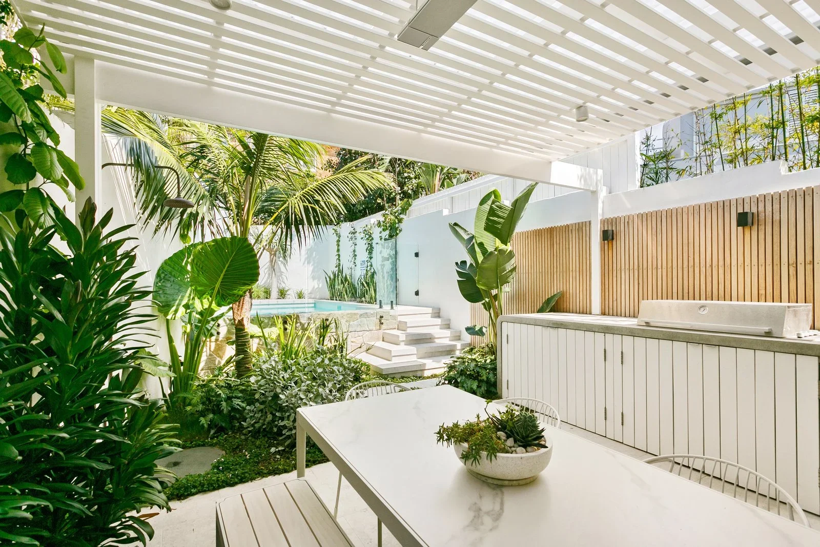 Outdoor patio with white table and chairs, lush tropical plants, a swimming pool, and a wooden privacy fence.