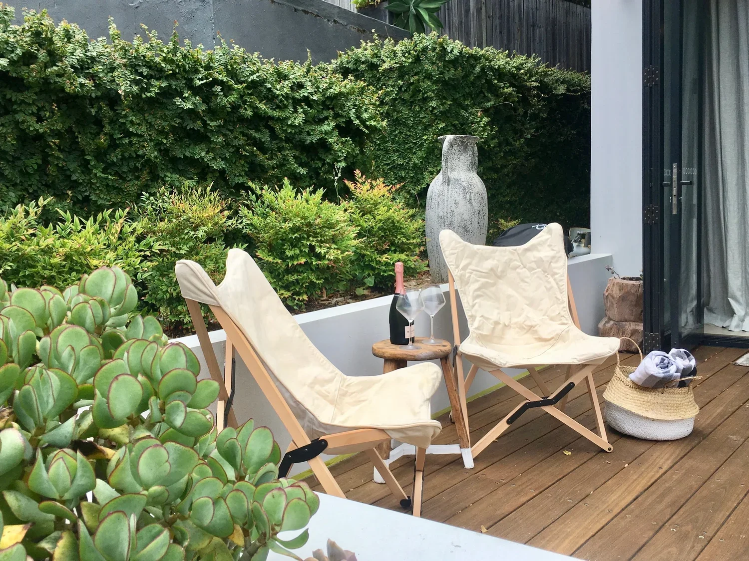 Outdoor patio with two foldable chairs, a small wooden table with a bottle of wine and two wine glasses, a woven basket with cloth, potted plants, and green shrubbery in the background.