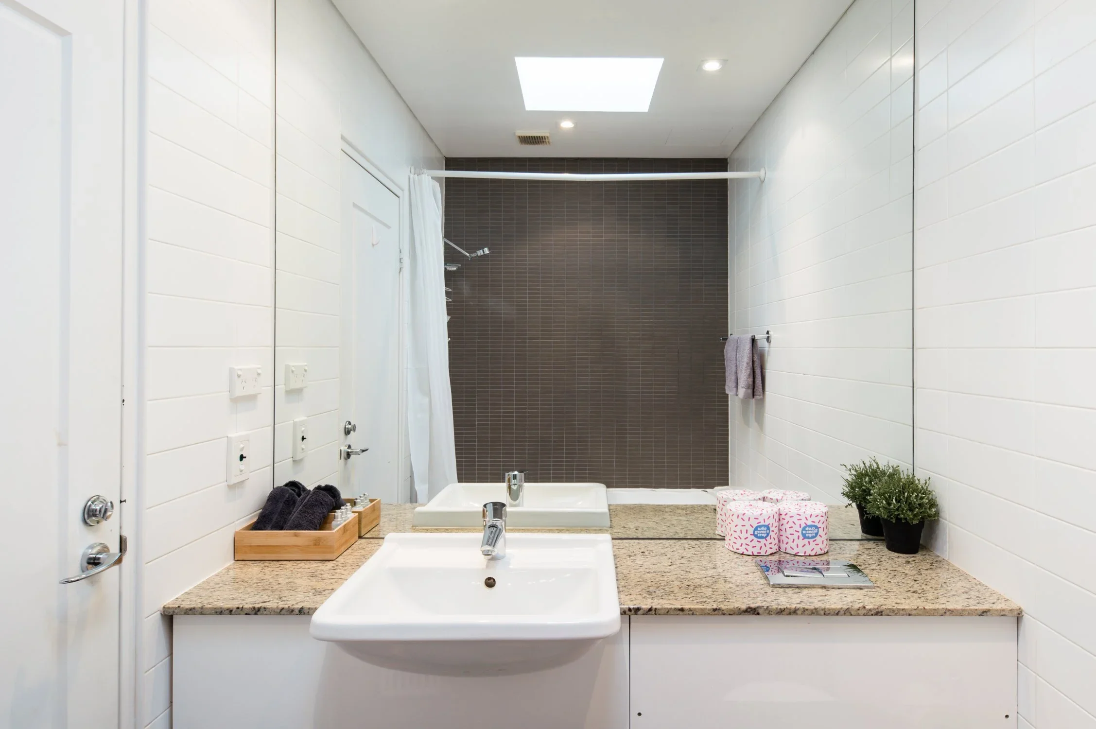 Bathroom with white tiled walls, granite countertop with double sink, mirror, a towel on the wall, a small potted plant, toiletries, and a shower area with a brown tiled wall and white shower curtain.