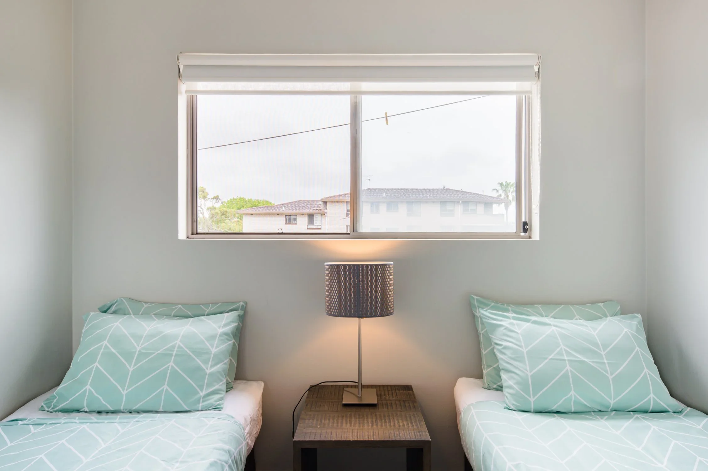 Two twin beds with matching light green and white geometric bedding, separated by a small wooden nightstand with a table lamp, in front of a window with a white blind.