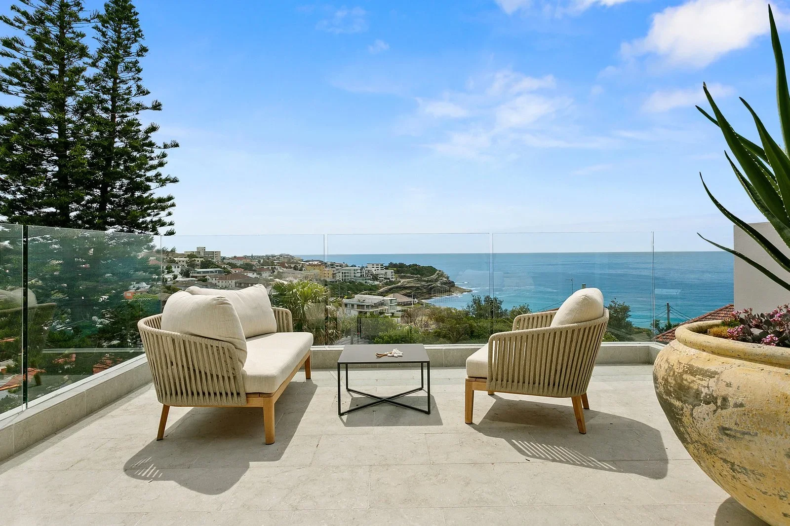 Rooftop patio with two beige cushioned lounge chairs and a small black coffee table, overlooking a coastal town and ocean under a partly cloudy sky.