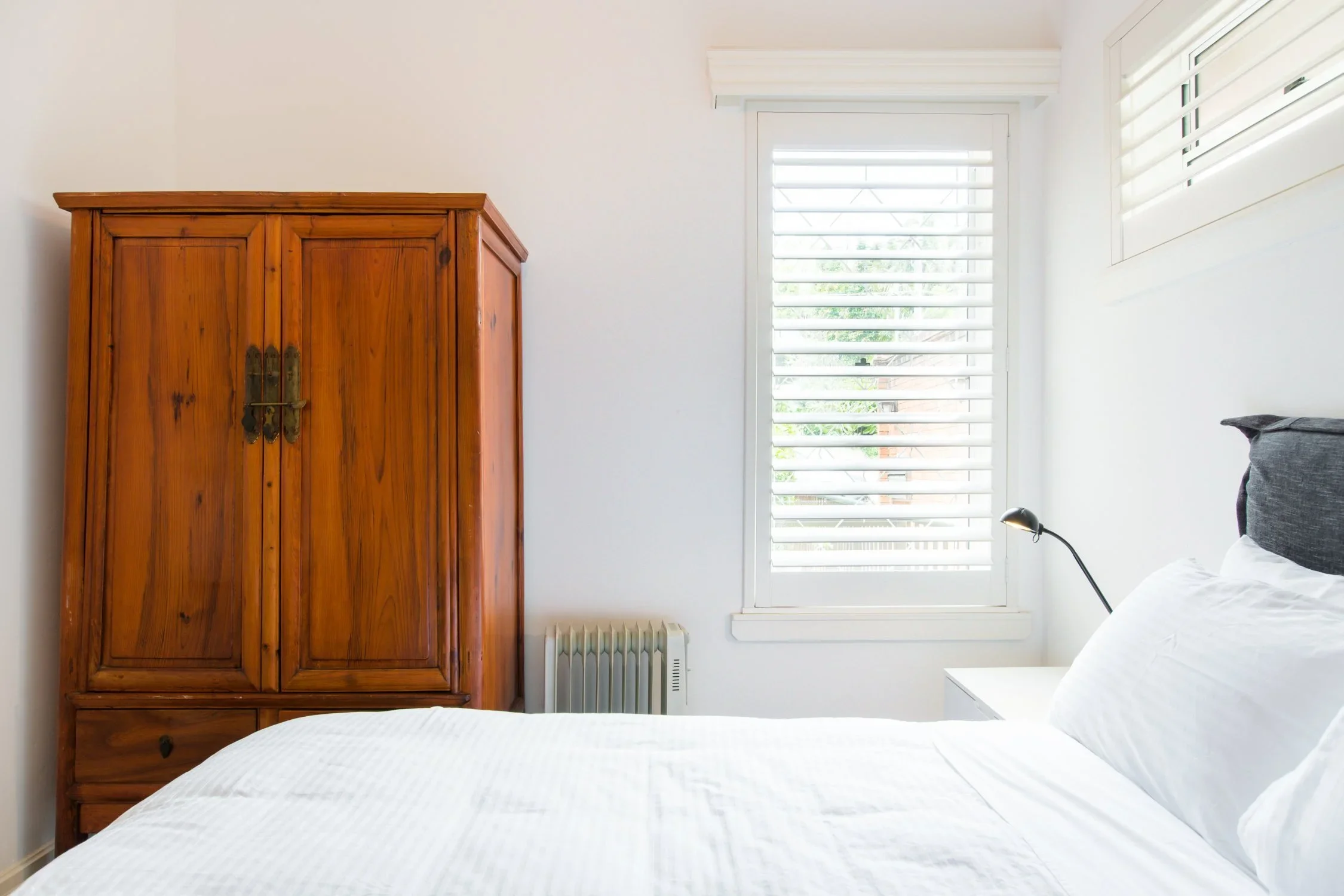 A bedroom with a white bed, a wooden wardrobe, a window with white blinds, a white nightstand, and a black reading lamp.