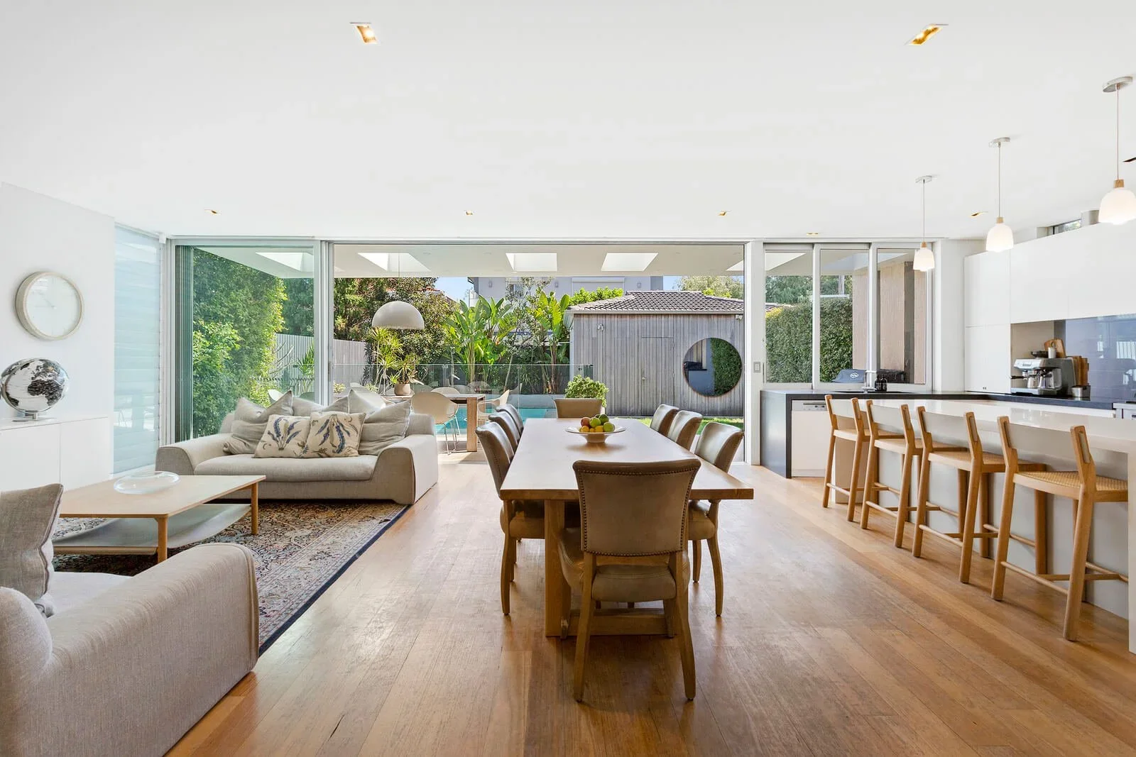 Bright open-plan living and dining area with large glass sliding doors, views of a backyard with trees, a patio, and a wooden shed, featuring wooden flooring, a white sofa with pillows, a dining table with chairs, a kitchen with a bar and stools, and