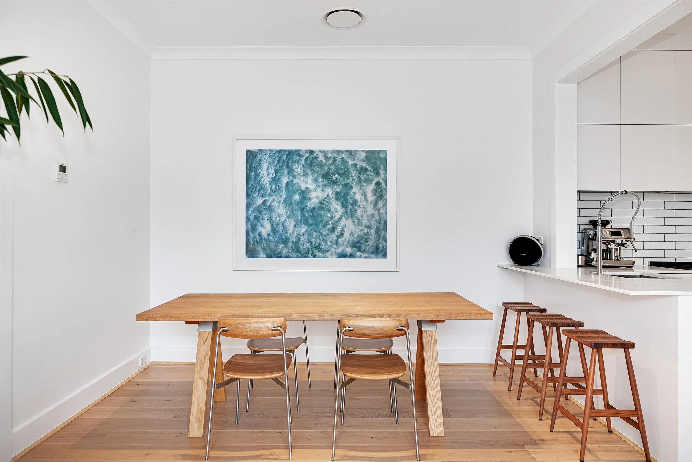 A dining area with a wooden table and four matching chairs, a framed ocean or water-themed artwork on the white wall, and bar stools at a kitchen counter with a coffee machine and black speaker.
