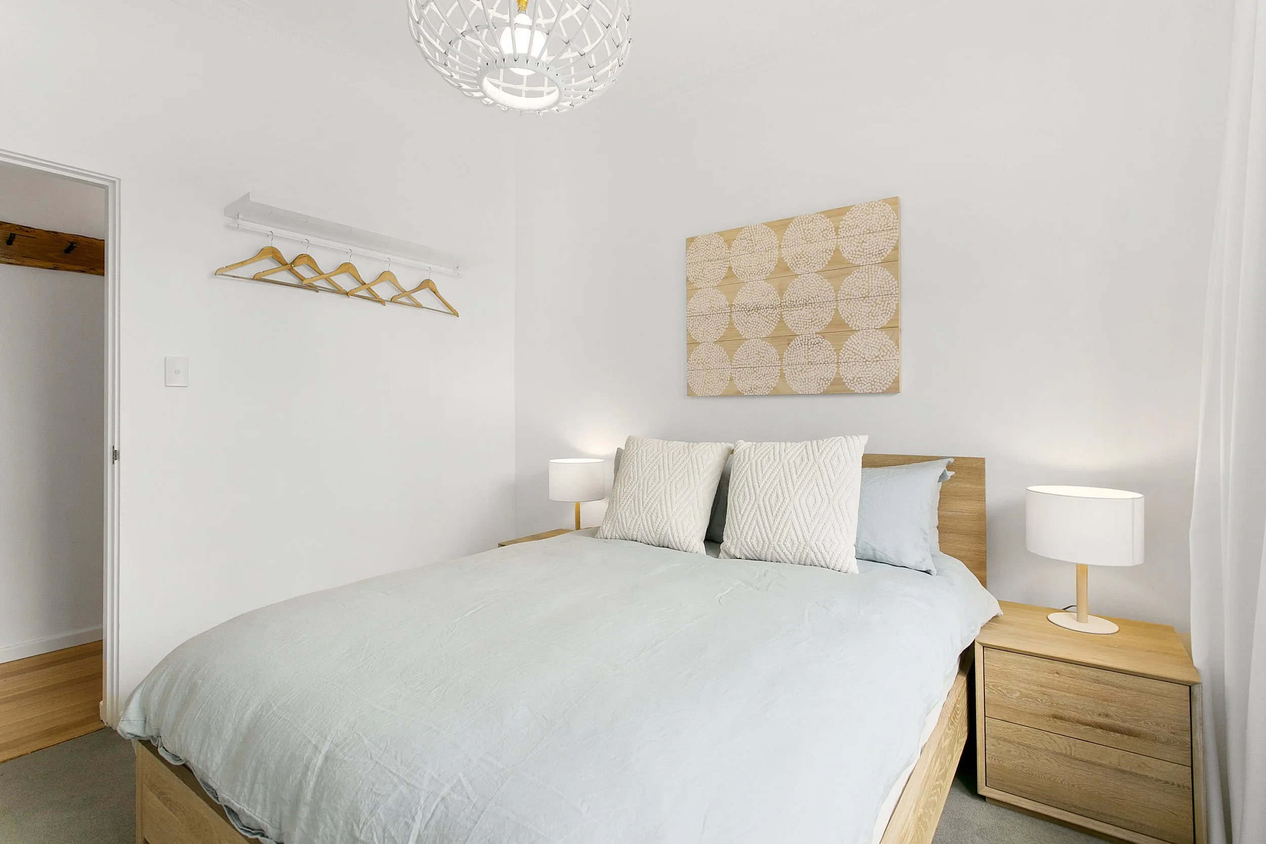 A minimalist bedroom with a wooden bed frame, white bedding, two white pillows, and a small wooden nightstand with a white lamp. There is a piece of abstract art above the bed and a wall-mounted coat rack with four empty wooden hangers.