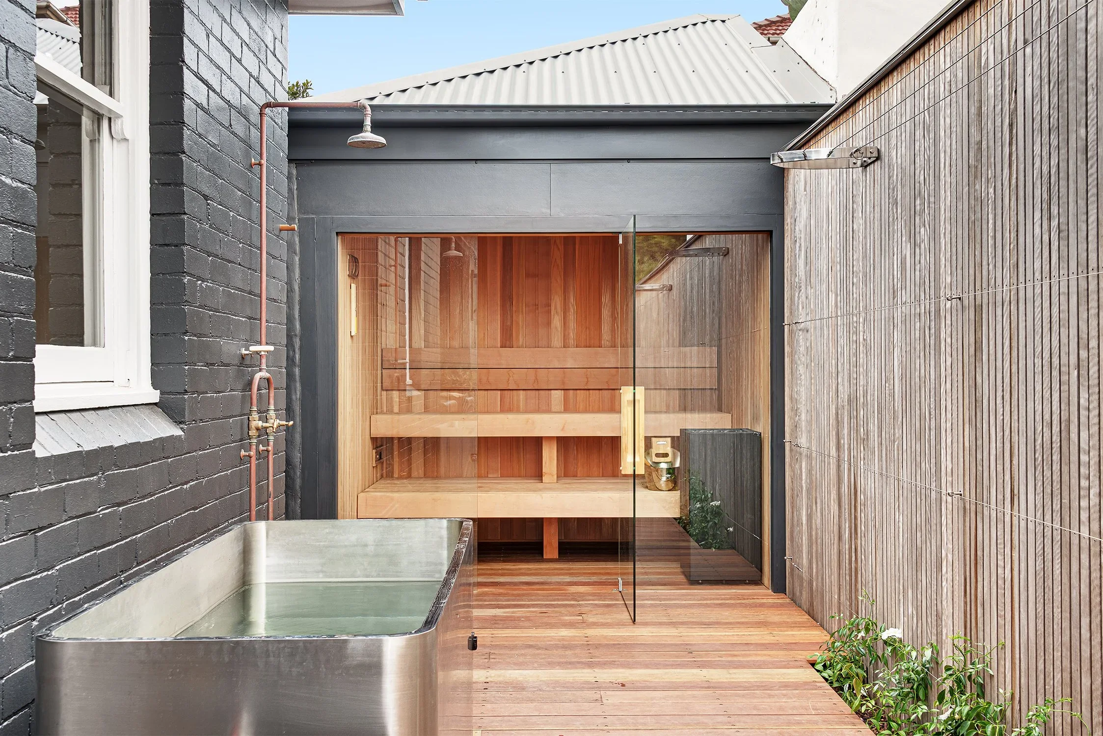 An outdoor sauna with glass doors situated between a black brick wall and a wooden fence. The sauna features wooden interior benches and is accessible via a sliding glass door. To the left, there is a metal sink with water, and a small plant is visib