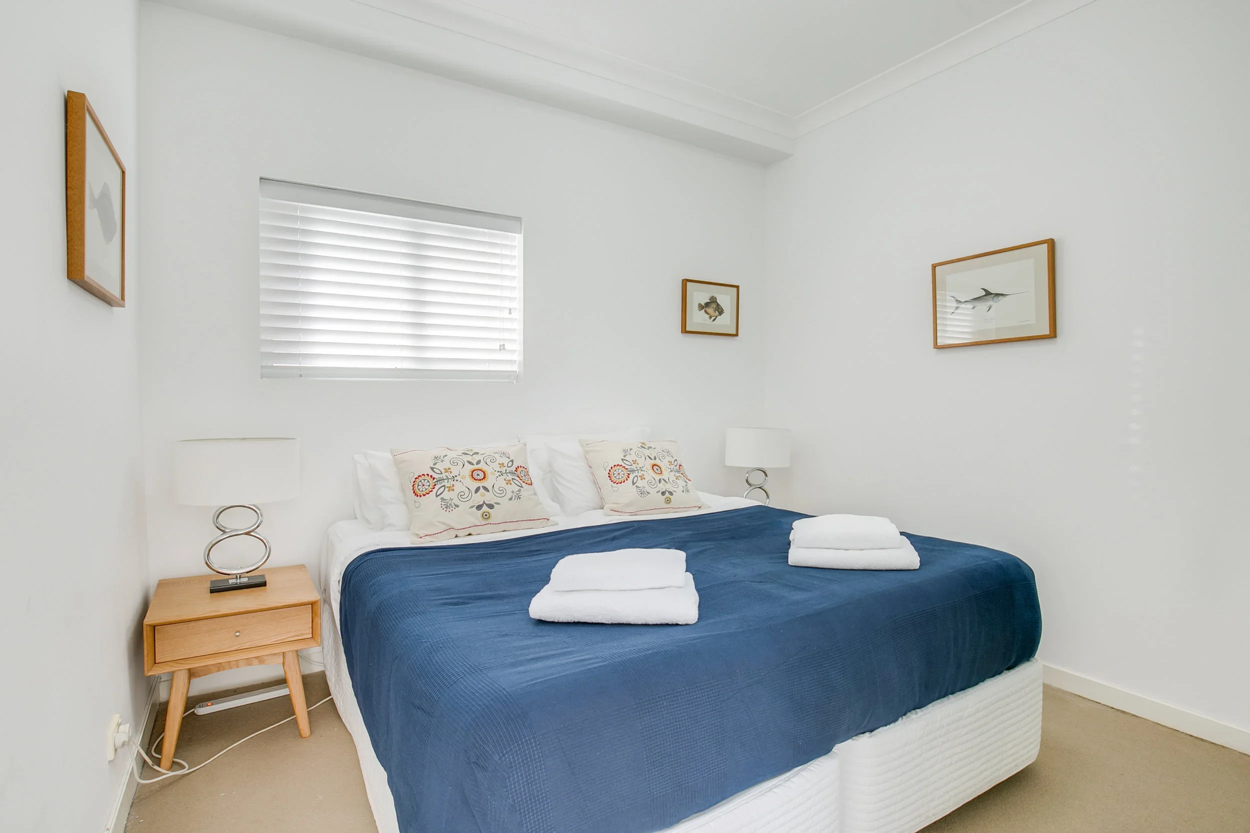 Clean and minimal bedroom with white walls, a window with blinds, a bed with white pillows and a navy blue blanket, two neatly folded towels on the bed, and framed fish artwork on the walls.