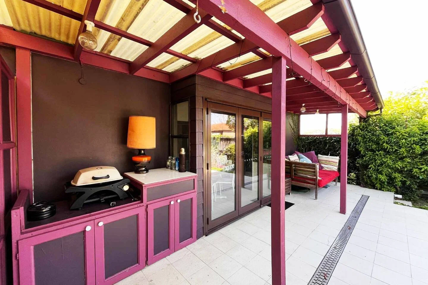 Outdoor patio with purple and black cabinets, a catering grill, a lamp, sliding glass doors, a purple cushioned seating area, and greenery on the sides.