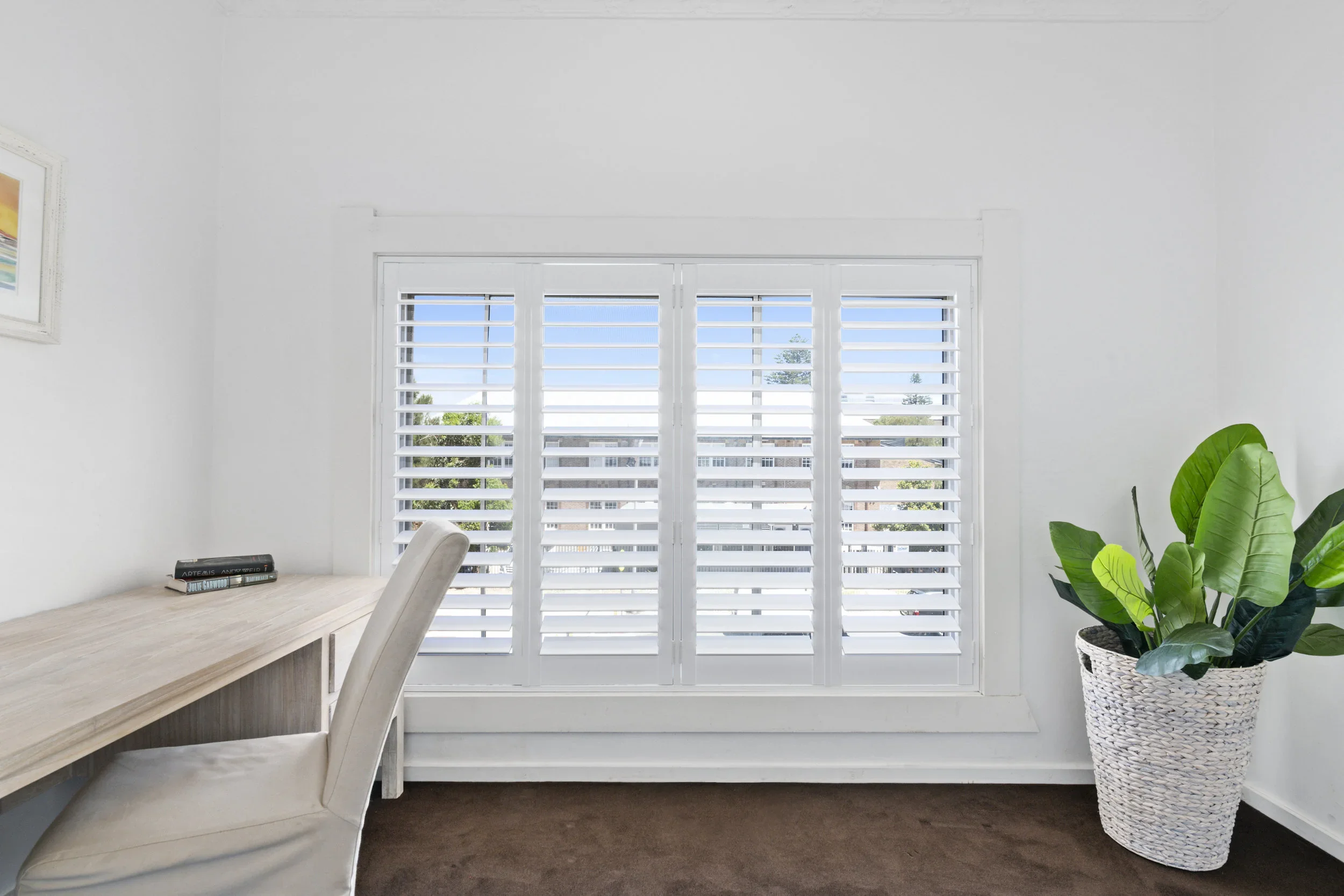 Room with a large window with white plantation shutters, a light-colored wooden desk with books, a white upholstered chair, a framed scenic picture on the wall, and a potted plant with large green leaves in a woven white basket.