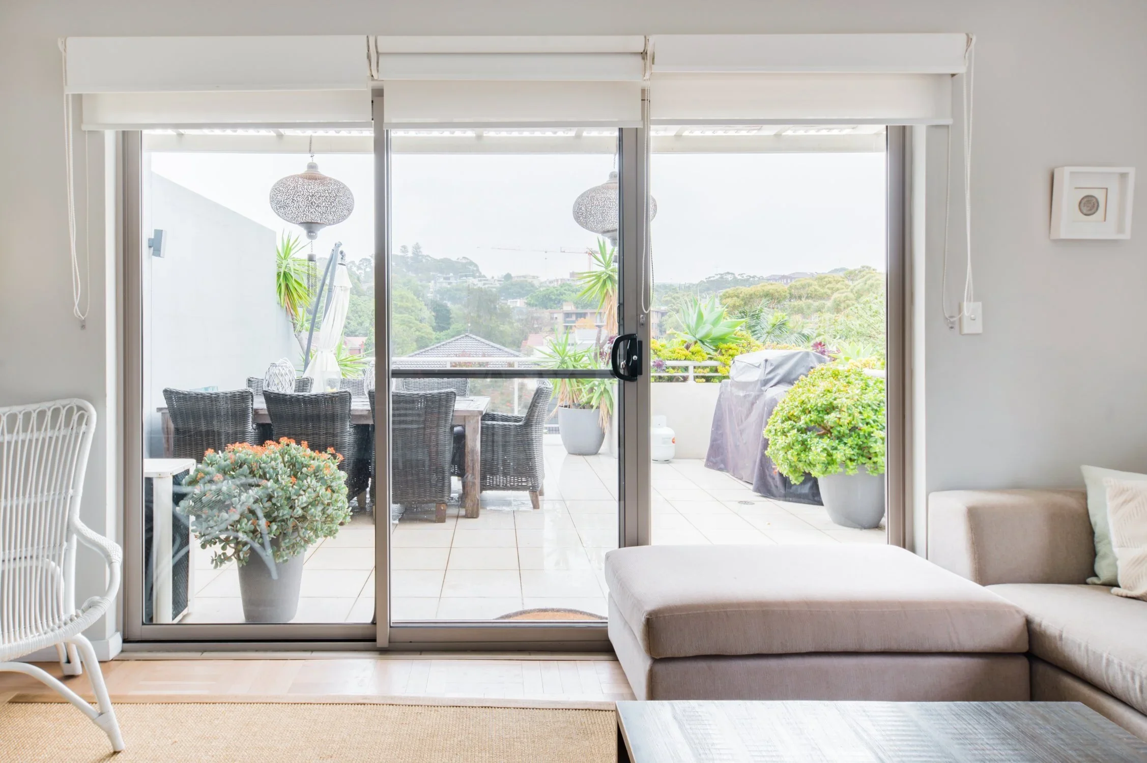 Living room view of sliding glass door leading to a balcony with outdoor dining table and potted plants, with a neighbor's roof and trees in the background.