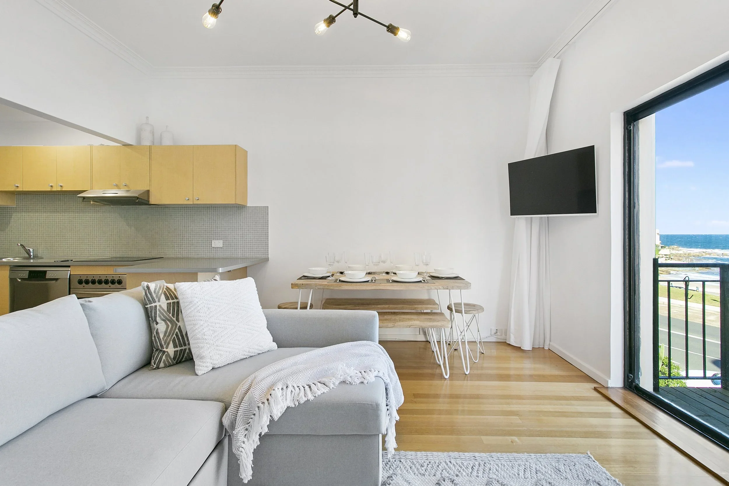Bright living room and dining area with white walls, a light gray couch with decorative pillows, a wooden dining table set with plates and glasses, and a large sliding glass door leading to a balcony overlooking the ocean.