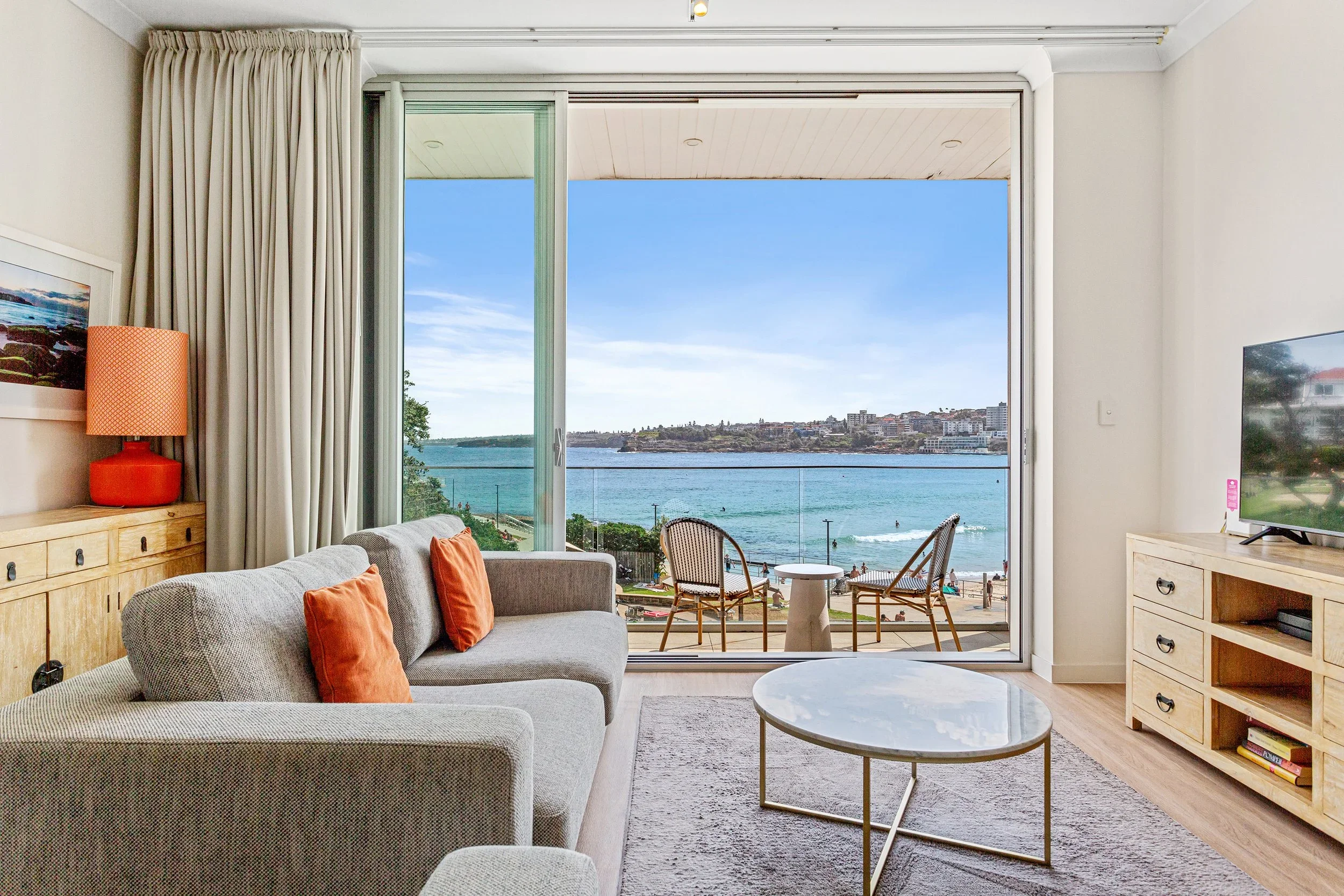 Living room with a view of the beach and water, sofa with orange pillows, wooden furniture, and chairs on the balcony.