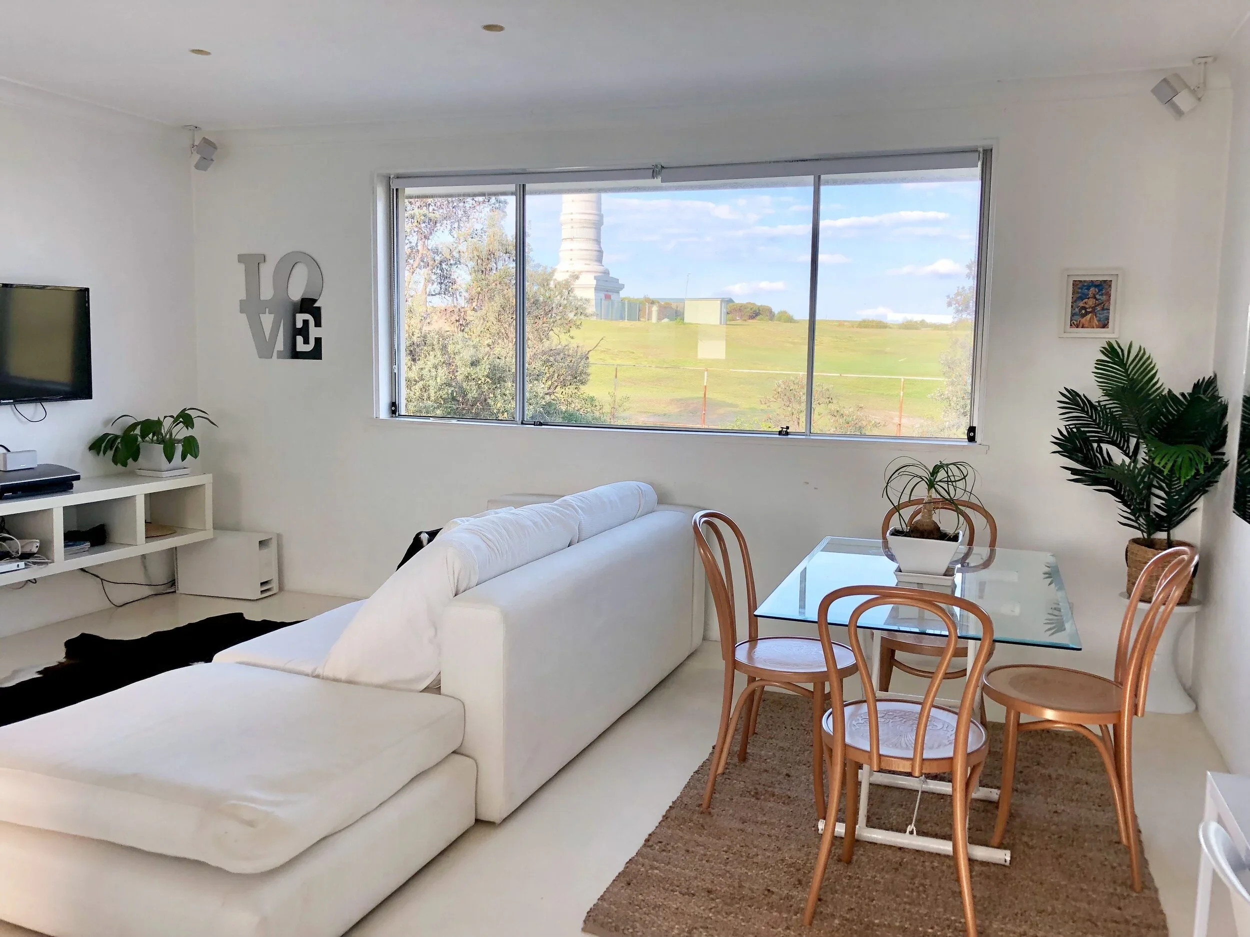 Living room with white sectional sofa, glass dining table with four wooden chairs, plants, and a large window showing an outdoor landscape with a monument or tower.