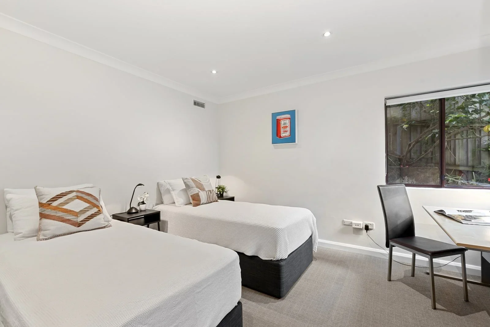 A bedroom with two white beds, each with decorative pillows, a black nightstand with a lamp and plant, a window showing trees outside, and a wall art piece of a fire alarm box. Work space with desk and chair - overlooking the garden area