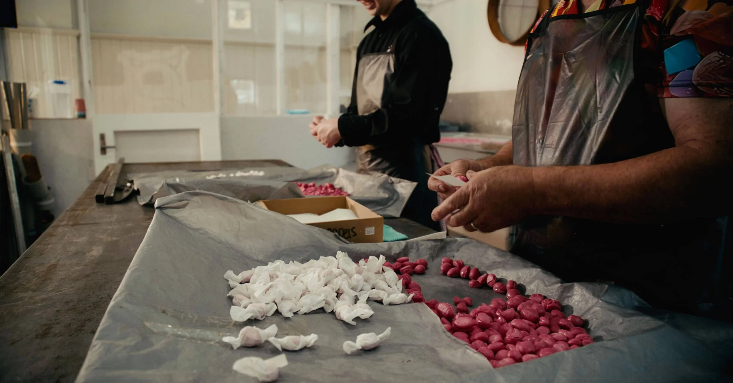 People working at a table sorting and packaging pink candies with white paper covers, in a room with white walls and a window.