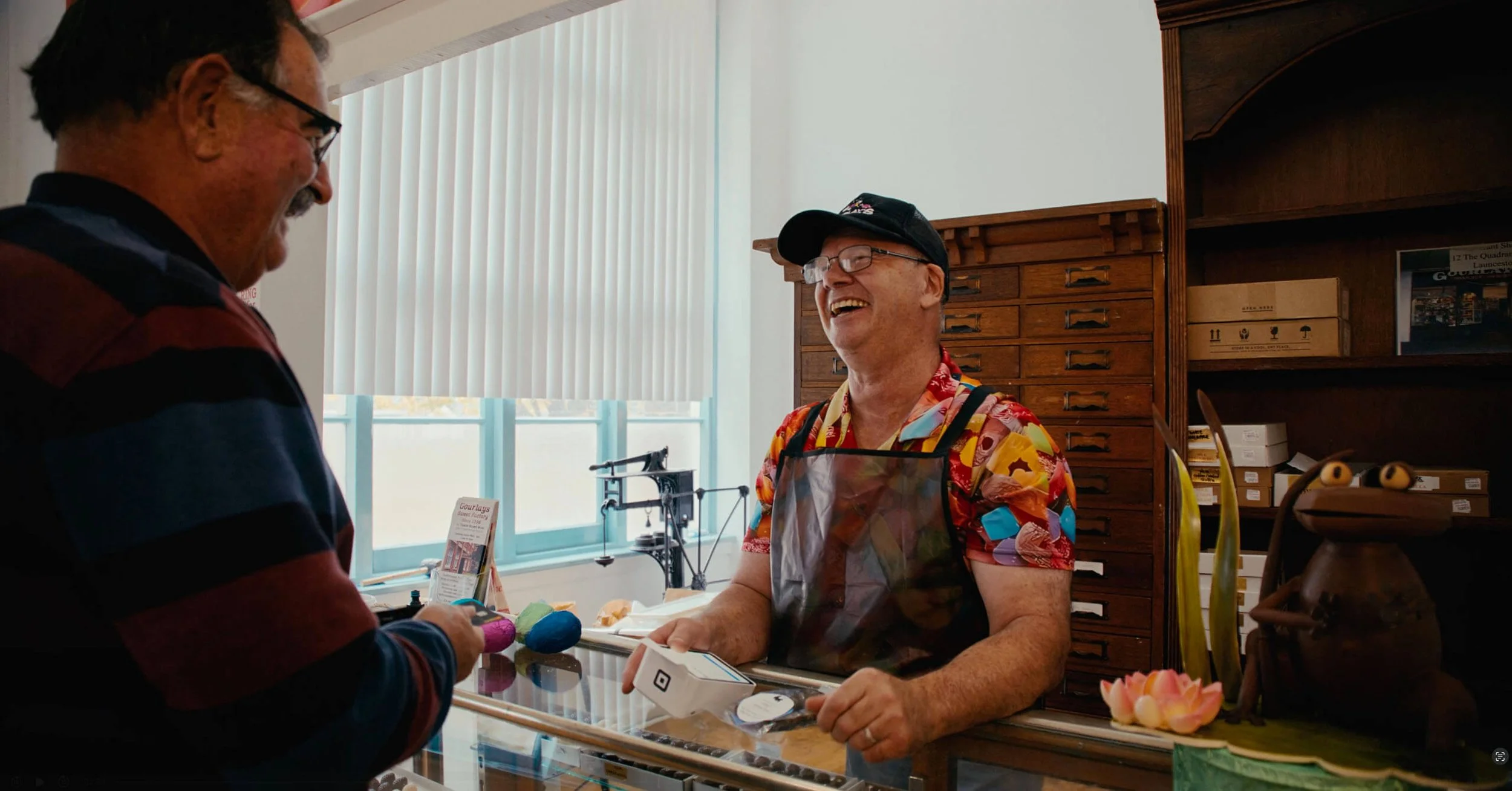 Two men at a jewelry counter smiling and chatting, with one man behind the counter wearing a colorful shirt and apron, and the other man wearing glasses and a striped shirt, holding a purple item.