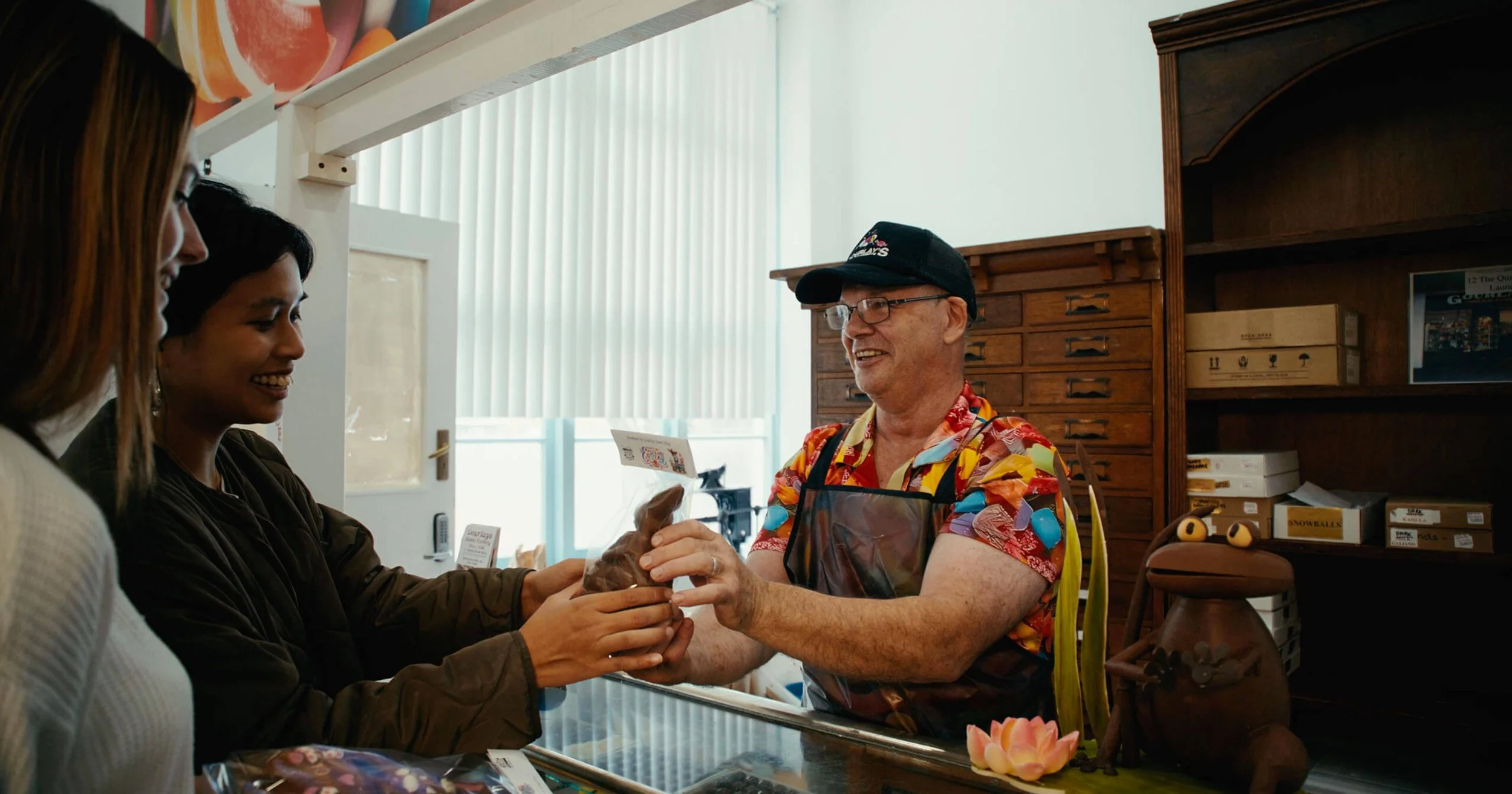 A man in a colorful Hawaiian shirt and apron hands a bag of food to two women at a counter inside a shop, smiling and engaging in conversation.
